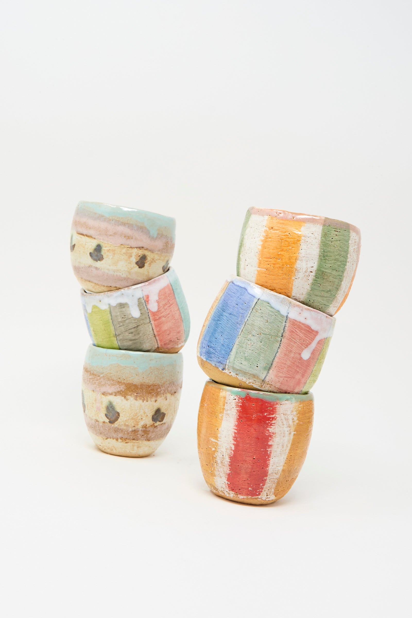 Six small, round Cup in Stripe ceramic cups by Shino Takeda are stacked in two uneven piles against a white background. Each hand-painted cup, made in New York, features stripes in pastel and earthy tones with abstract patterns.
