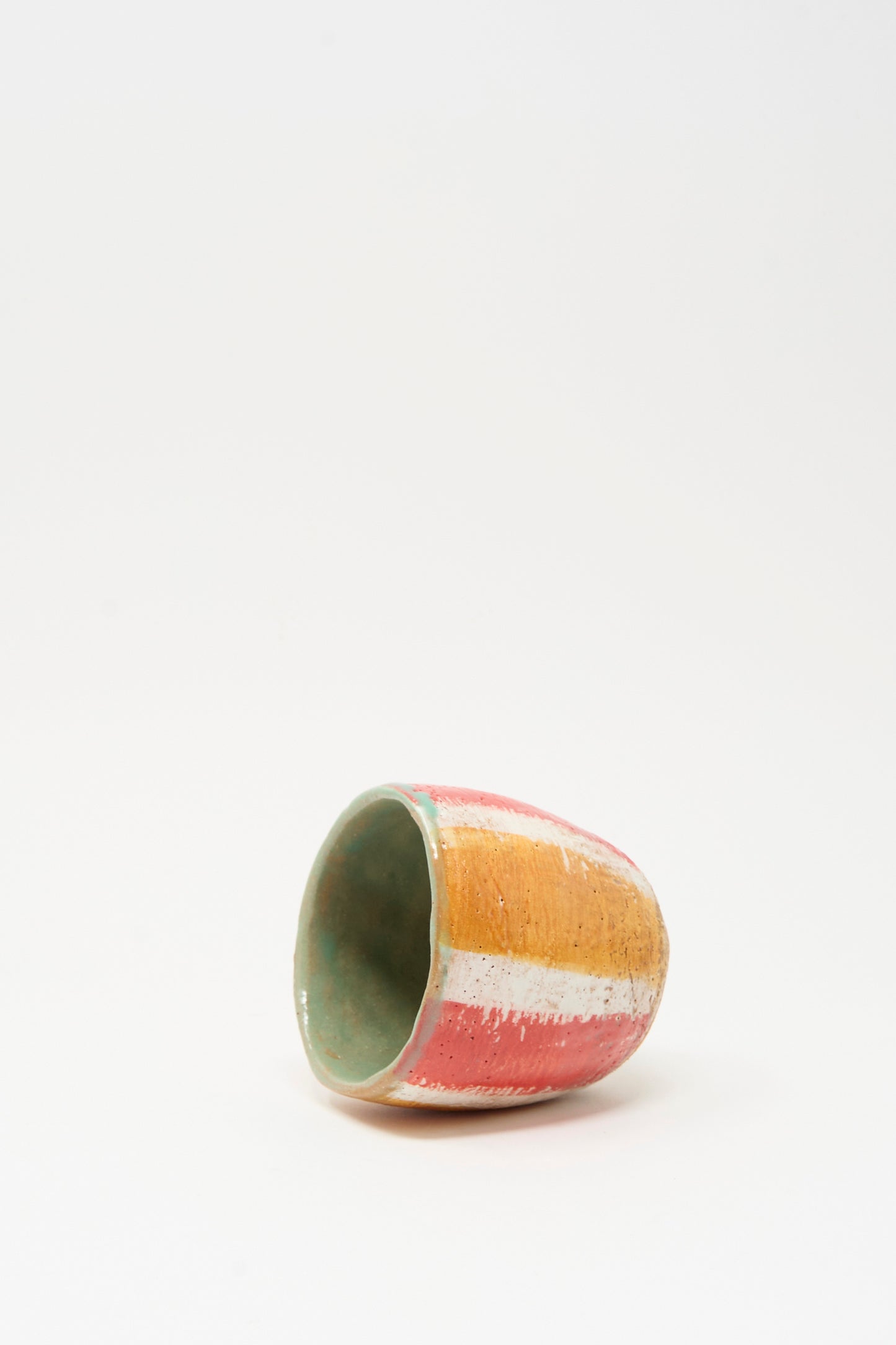 The Cup in Stripe II by Shino Takeda, a small hand-painted ceramic piece featuring orange, white, and pink stripes, rests on its side against a plain white background.