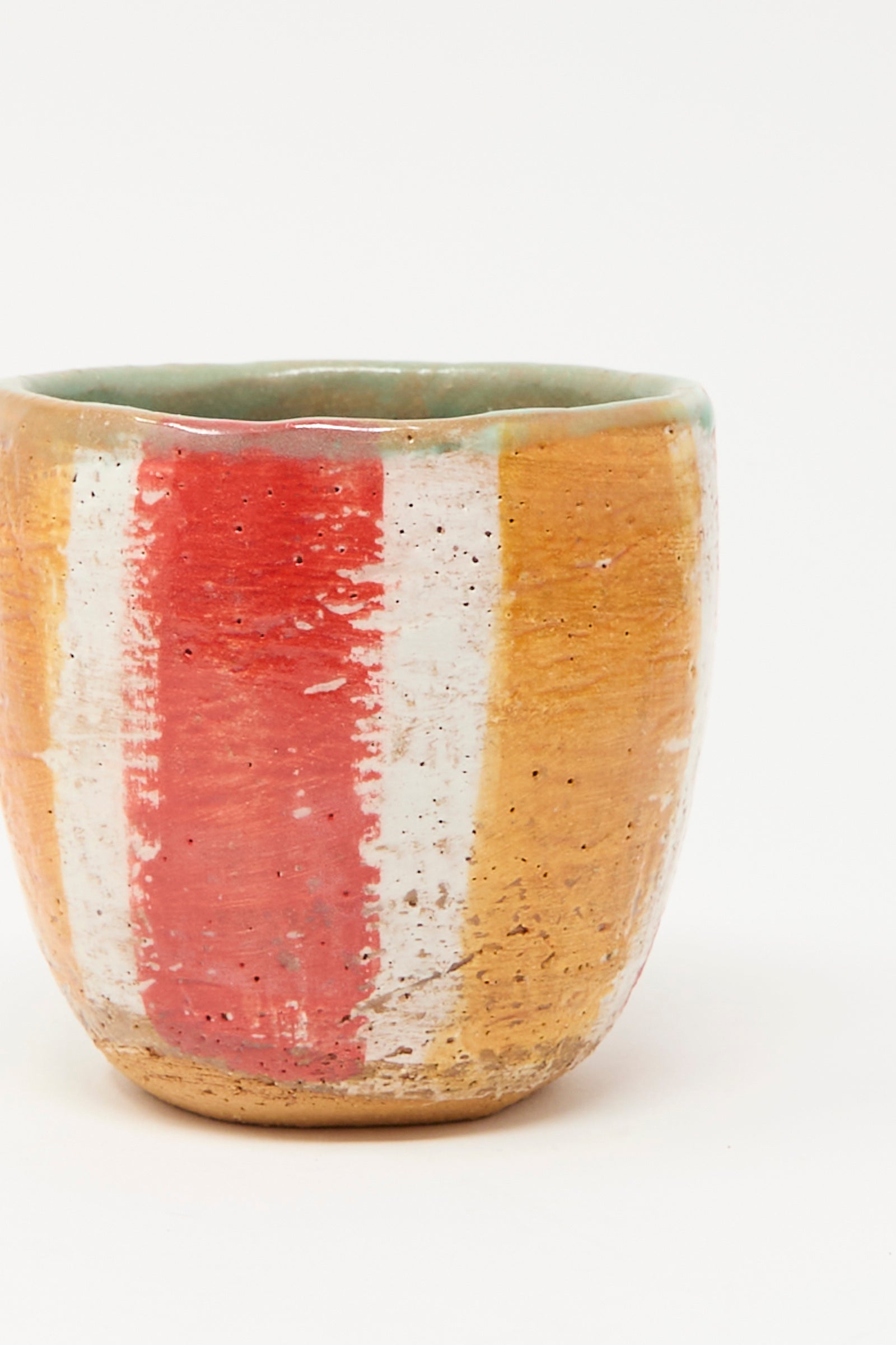 The Cup in Stripe II by Shino Takeda is a hand-painted ceramic cup with a rounded form and vertical red, white, and ochre stripes on a textured surface, shown against a plain white background.