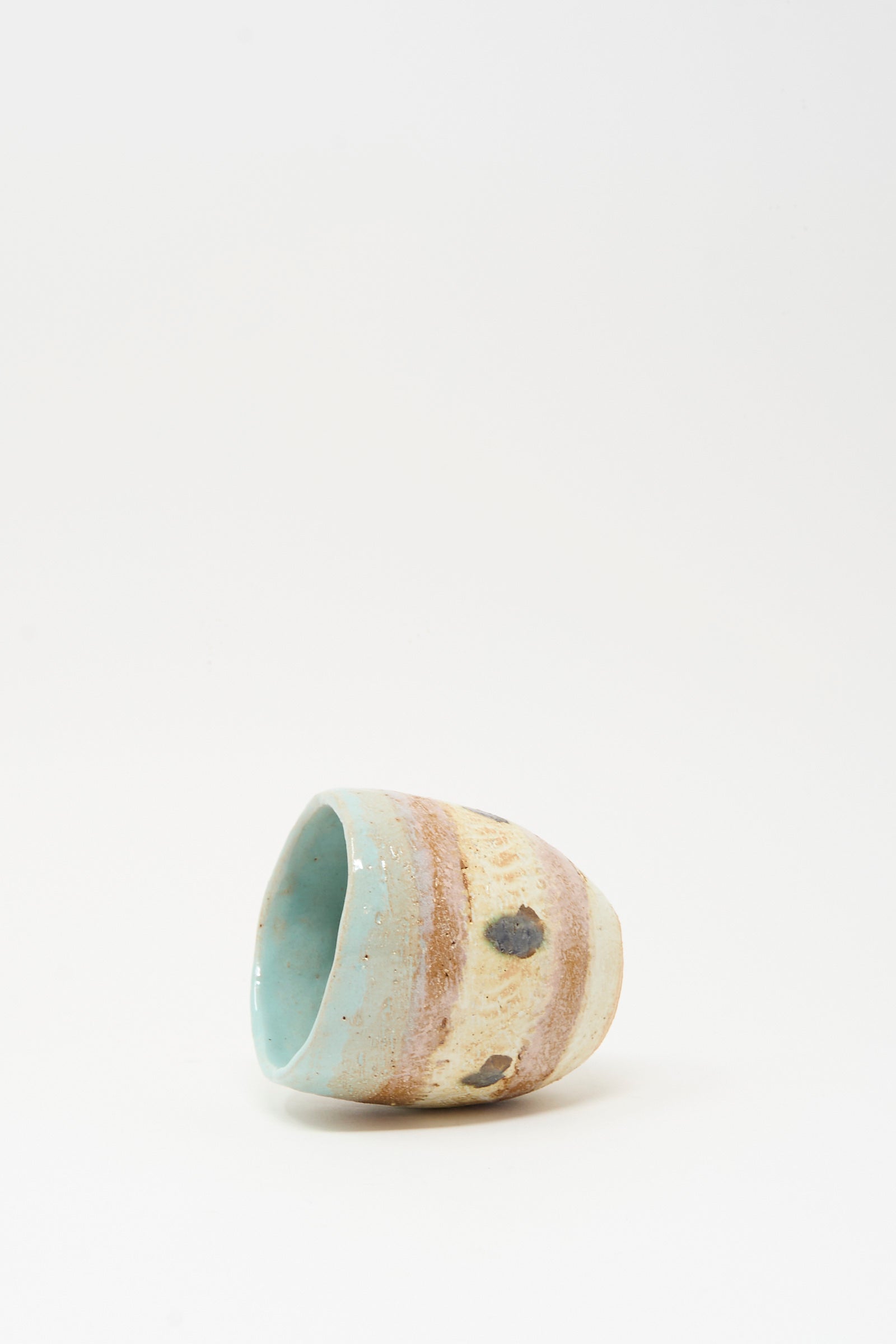 The Cup Tall in Dots II by Shino Takeda, a hand-painted ceramic with a blue interior and beige, brown, and blue-speckled exterior, lies on its side against a plain white background.