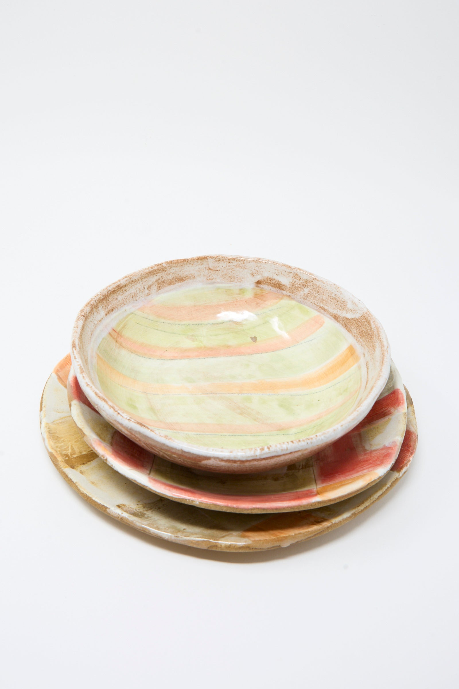 Three ceramic plates with hand-painted pastel patterns are stacked on a white background, next to the Serving Bowl in Stripes by Shino Takeda.
