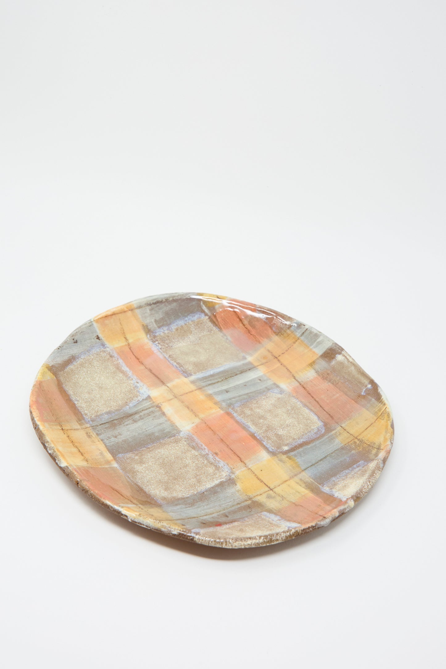 A round ceramic plate from Shino Takeda, hand-painted with a plaid pattern in pastel shades of orange, gray, and beige on a white background.