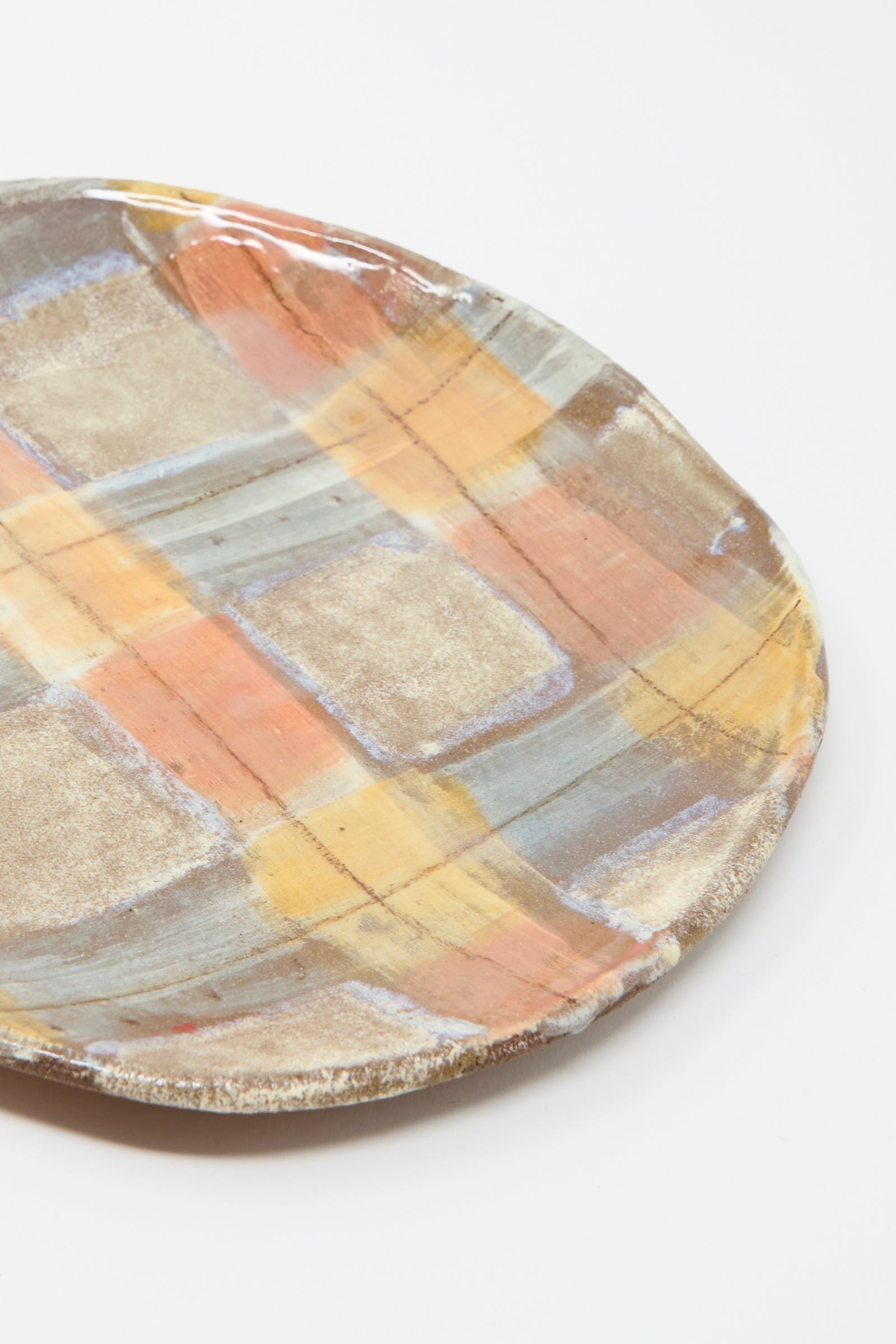 A closeup of the Shallow Plate in Pastel Plaid by Shino Takeda is a round clay plate with a hand-painted plaid pattern in soft hues of orange, brown, and gray on a vivid white background.