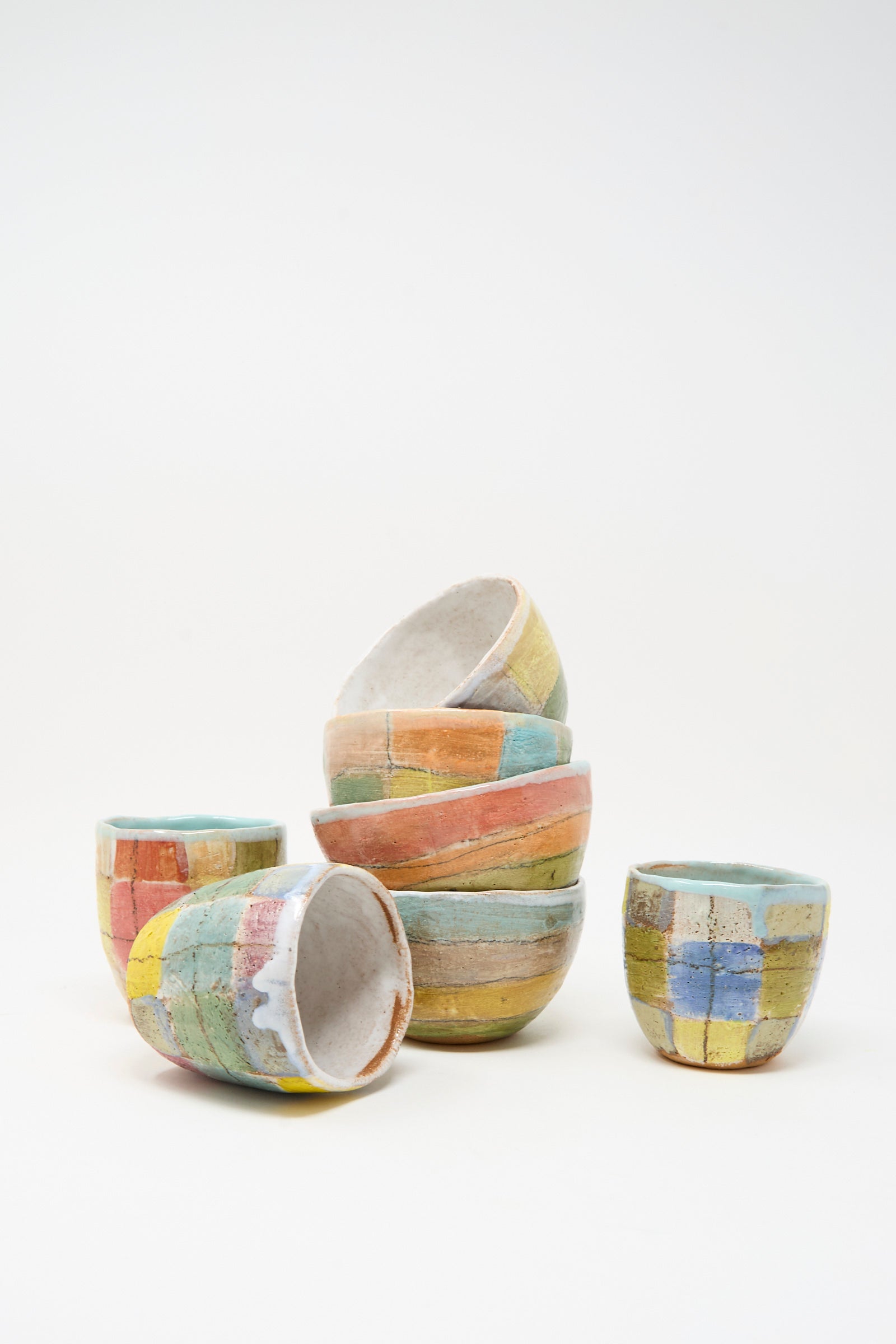 A stack of colorful, handmade ceramic cups and the Shino Takeda Small Bowl in Check Multi I with hand-painted geometric patterns, arranged against a plain white background.