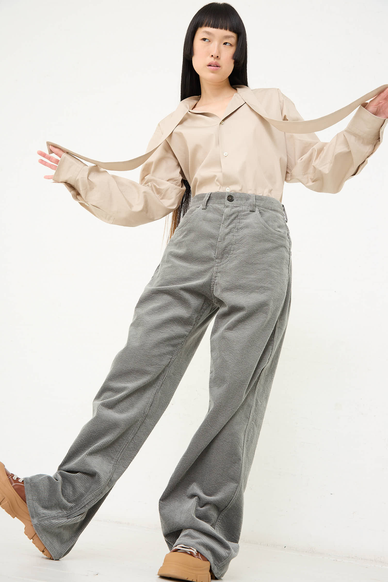 A person wearing a beige long-sleeve top with oversized sleeves and a relaxed fit pairs it with Sofie D'Hoore Cotton Corduroy Pavia Pants in Sage, posing against a plain white background.