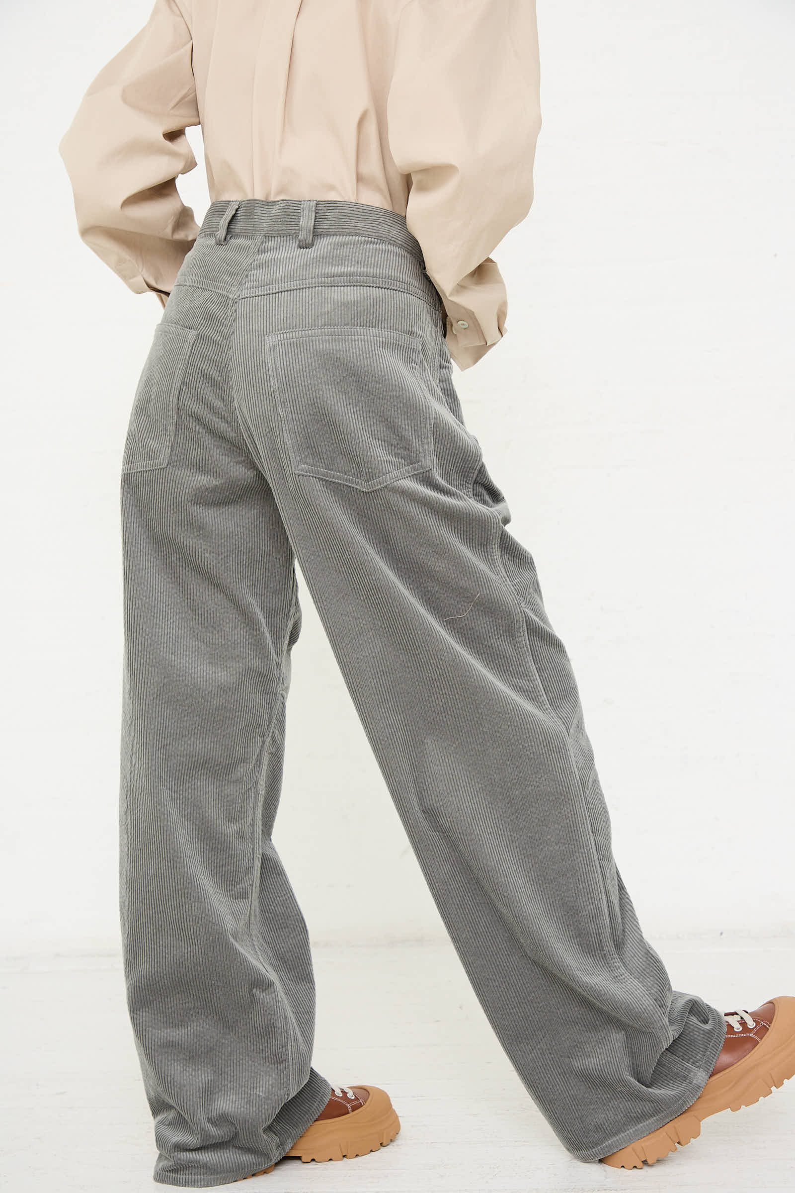 A person wearing a beige shirt and the Cotton Corduroy Pavia Pant in Sage by Sofie D'Hoore stands with hands on hips. The wide-legged, relaxed fit pants complement their brown shoes with thick soles.