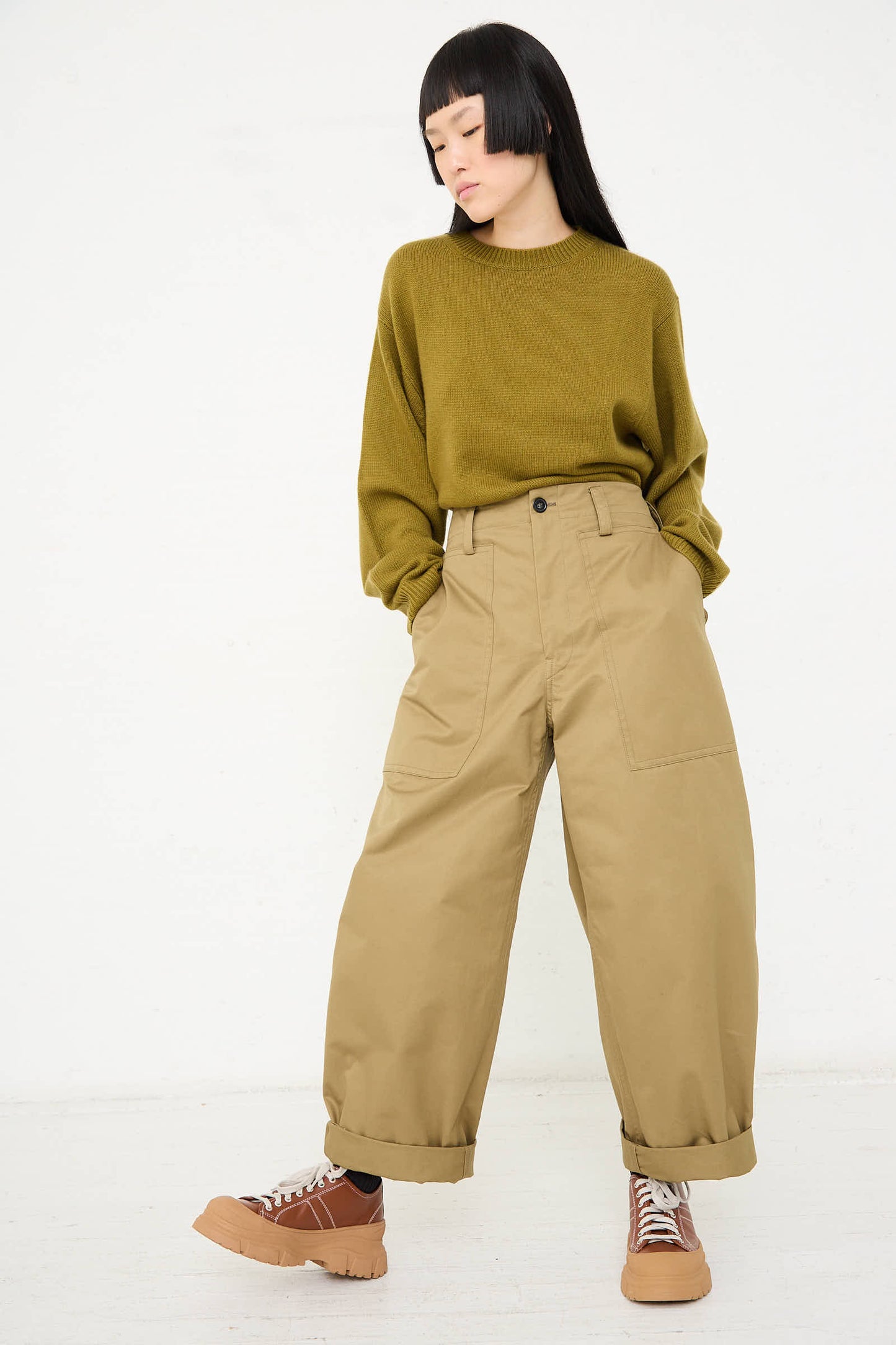 A person wearing a green sweater, Sofie D'Hoore's Cotton Gabardine Phoenix Pant in Pepper with large pockets, and brown shoes stands with hands in pockets against a plain white background.