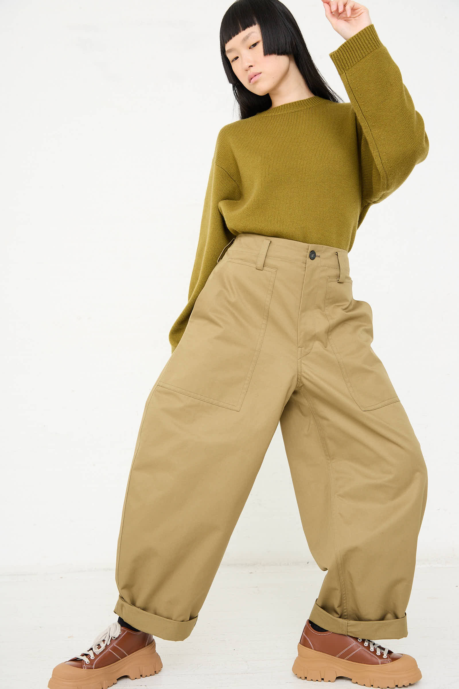 Person wearing an oversized green sweater and Sofie D'Hoore's Cotton Gabardine Phoenix Pant in Pepper, posing with one arm raised. They are also wearing brown shoes with thick soles.