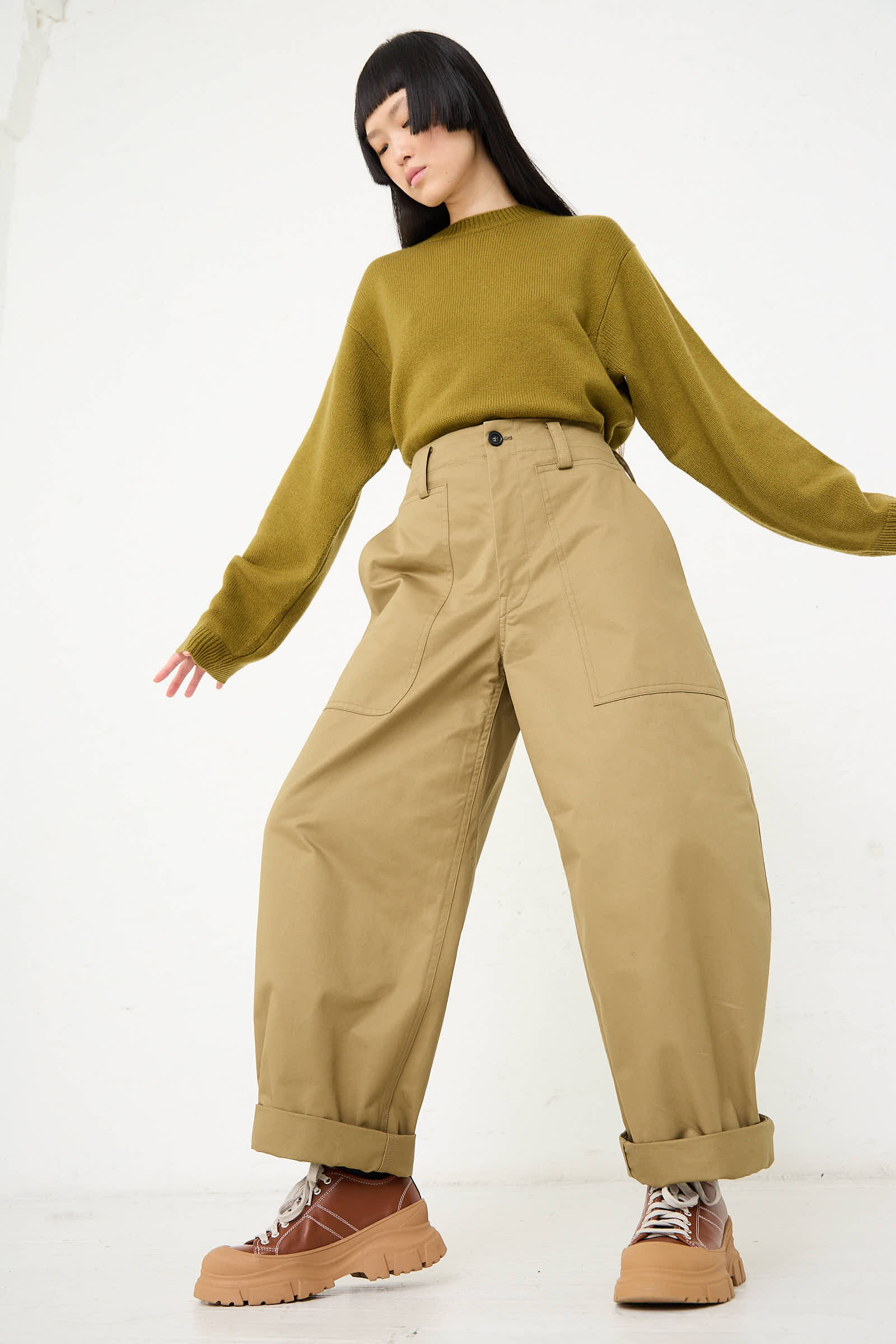 A person with long black hair is wearing a green sweater, the Cotton Gabardine Phoenix Pant in Pepper by Sofie D'Hoore, and tan shoes, standing against a white background.