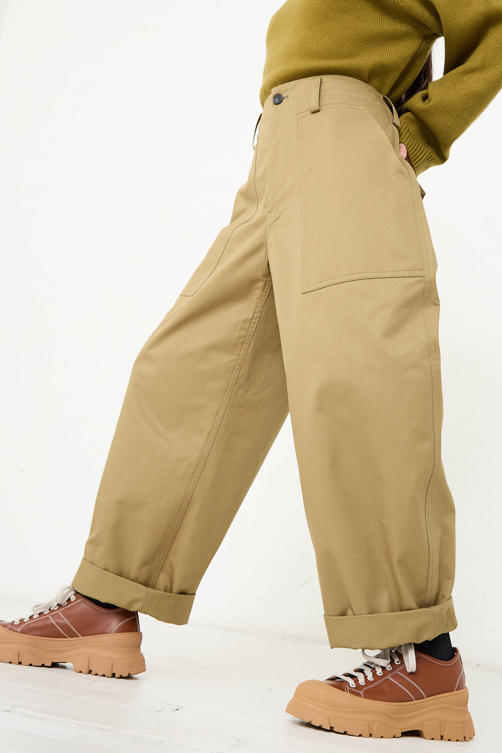 A person is wearing the Sofie D'Hoore Cotton Gabardine Phoenix Pant in Pepper, featuring beige wide-leg pants with an oversized fit, large pockets, and rolled cuffs. They have paired it with brown chunky-soled sneakers and a green sweater. The background is plain white.