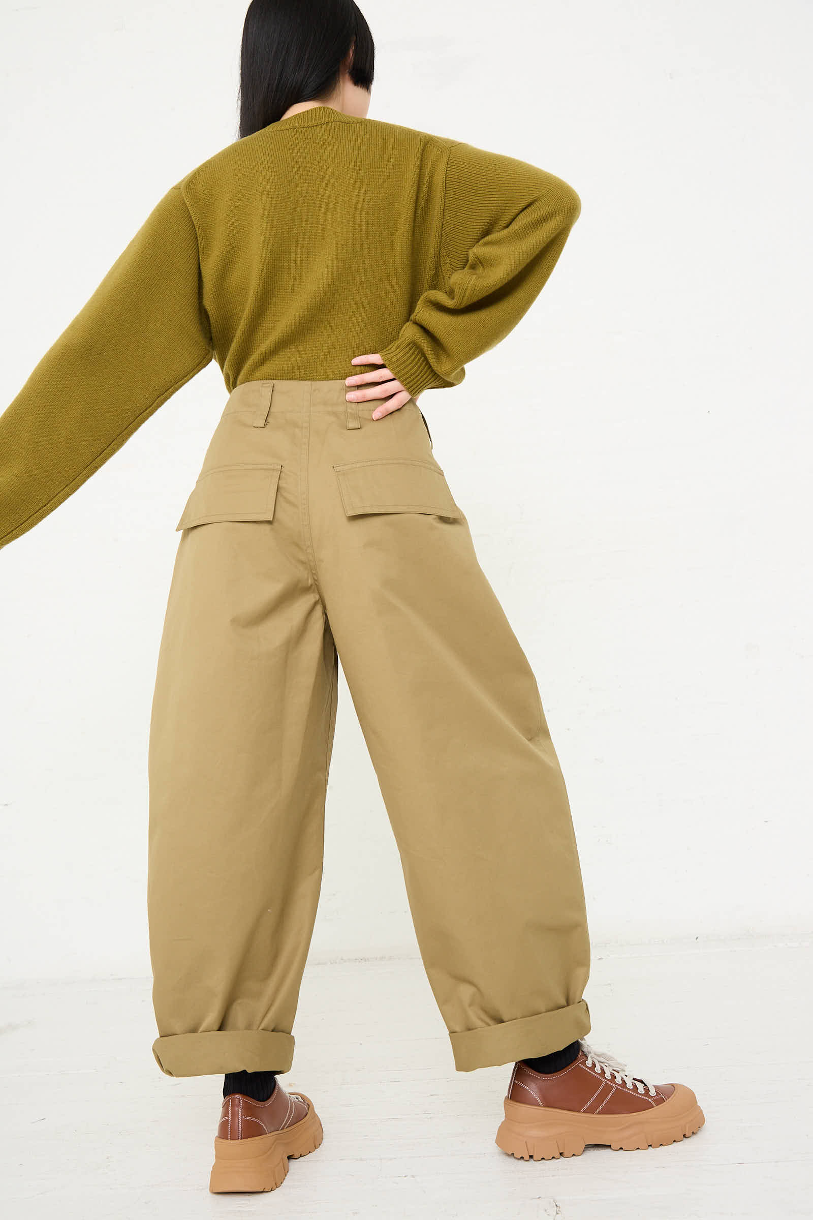 A person with long black hair is wearing a green sweater, Sofie D'Hoore Cotton Gabardine Phoenix Pant in Pepper, and brown shoes, posing with one hand on their hip and back turned to the camera.