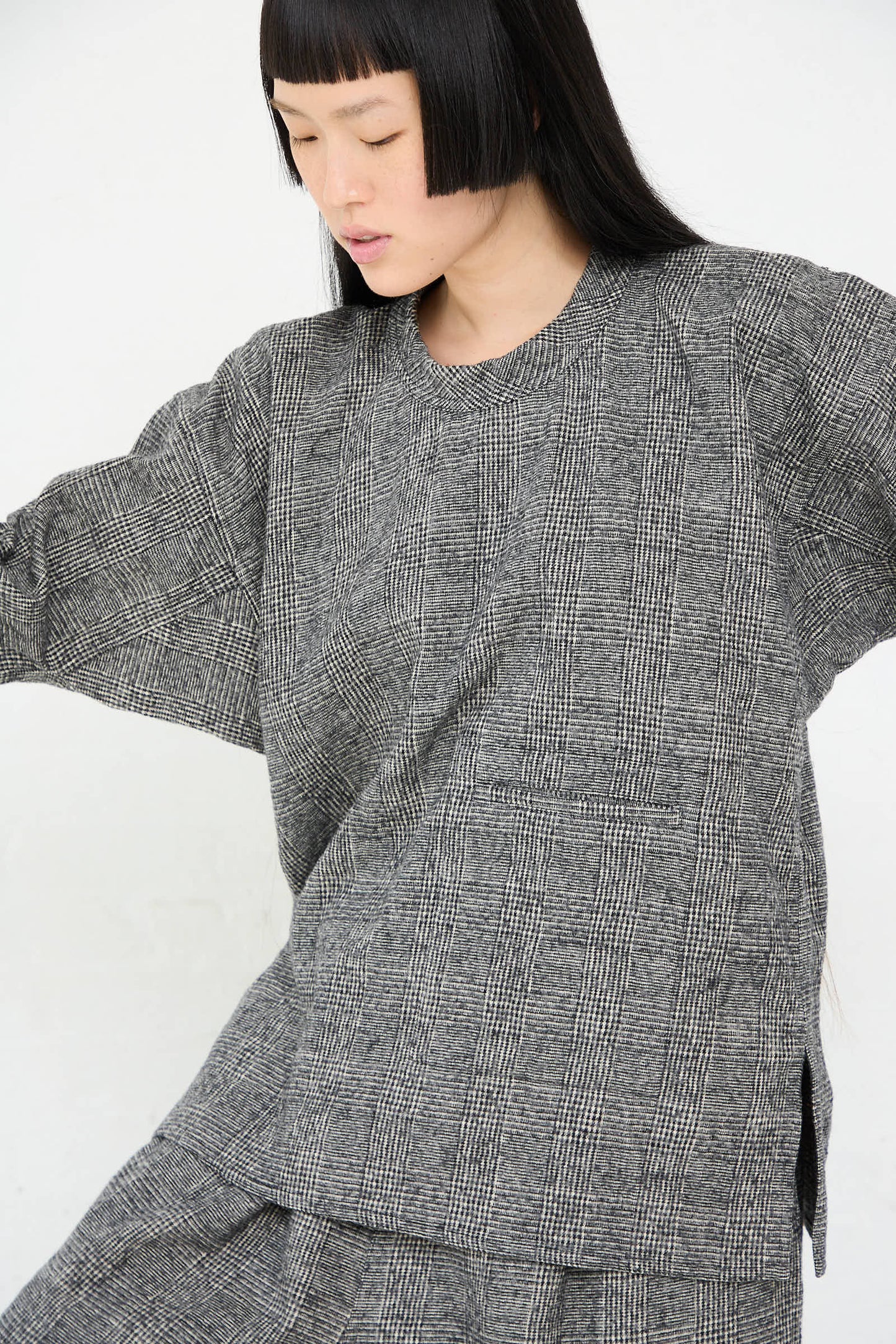 A person with straight black hair and bangs is wearing the Sofie D'Hoore Cotton Wool Washed Biba Top in Mid Grey. The individual appears to be moving or stretching, with their arms slightly raised, against a plain white background.