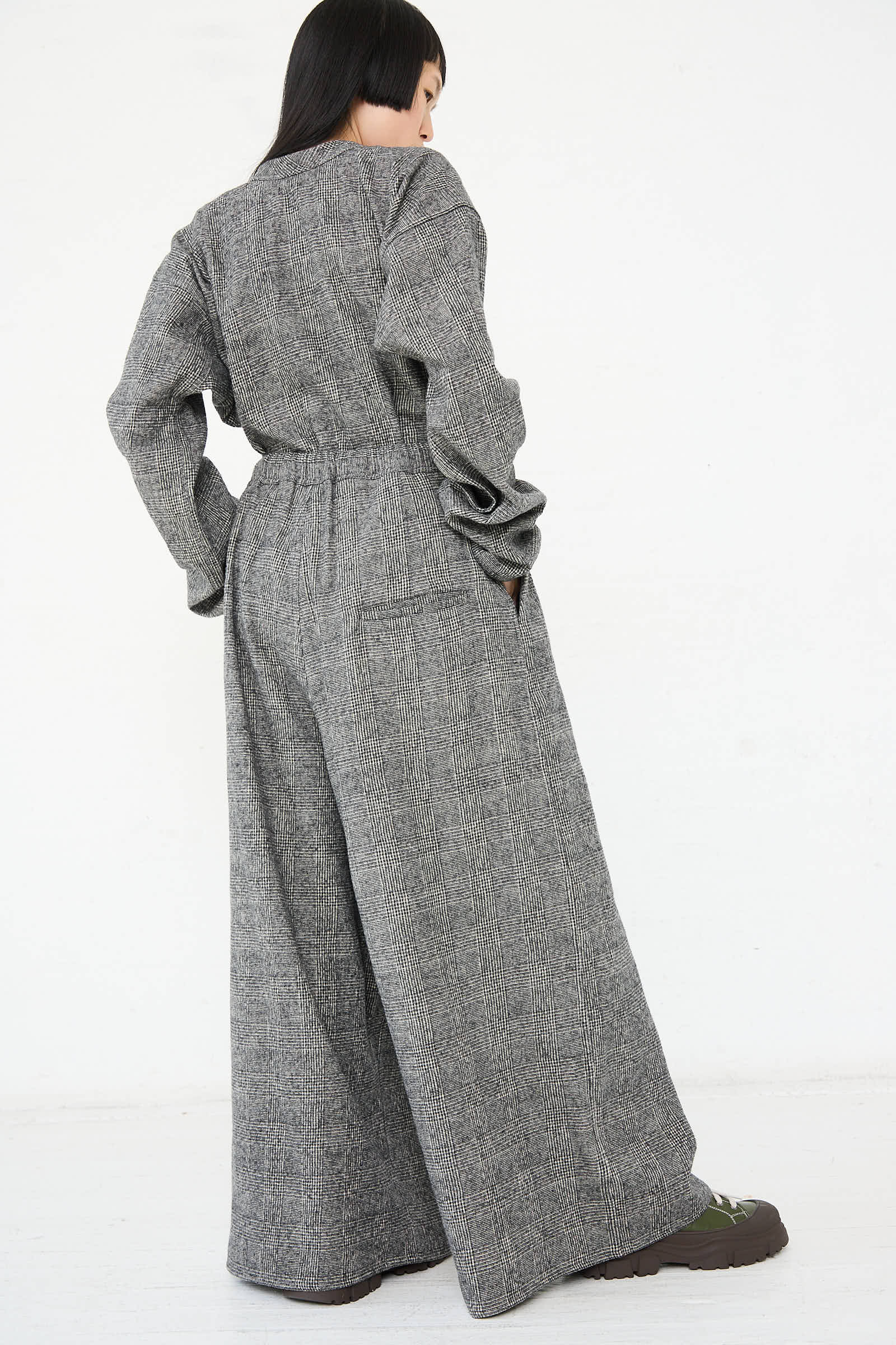 Person standing and facing away, wearing Sofie D'Hoore's Cotton Wool Washed Palau Pant in Mid Grey, a gray long-sleeve jumpsuit with wide legs reminiscent of wide-leg pants, paired with dark shoes.