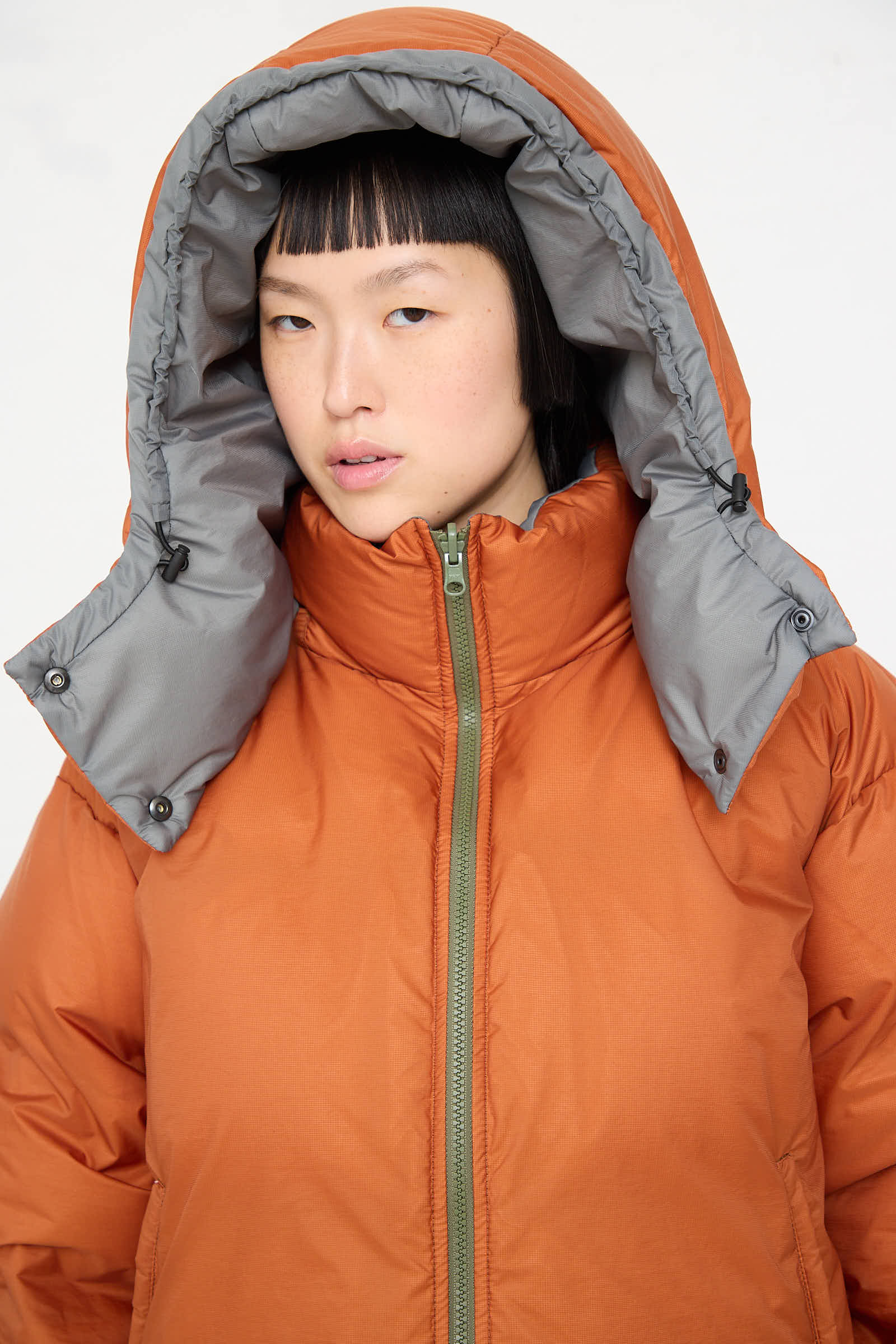 A person with short black hair wearing a Sofie D'Hoore Down Filled Odna Reversible Jacket in Rust and Sage stands against a plain background.