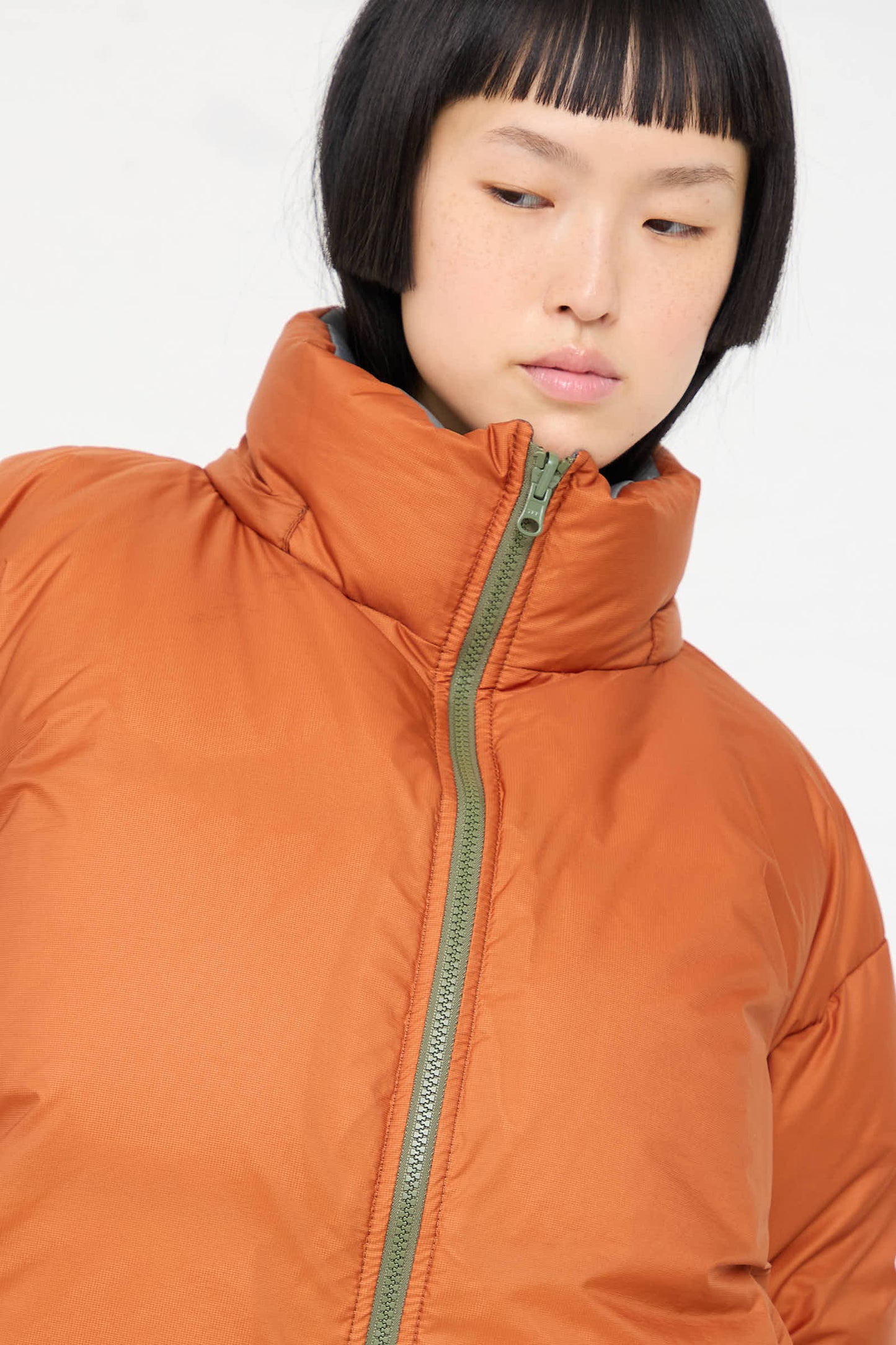 A person with short hair wearing a Sofie D'Hoore Down Filled Odna Reversible Jacket in Rust and Sage, featuring a high collar and front zipper, looking slightly down and to the side.