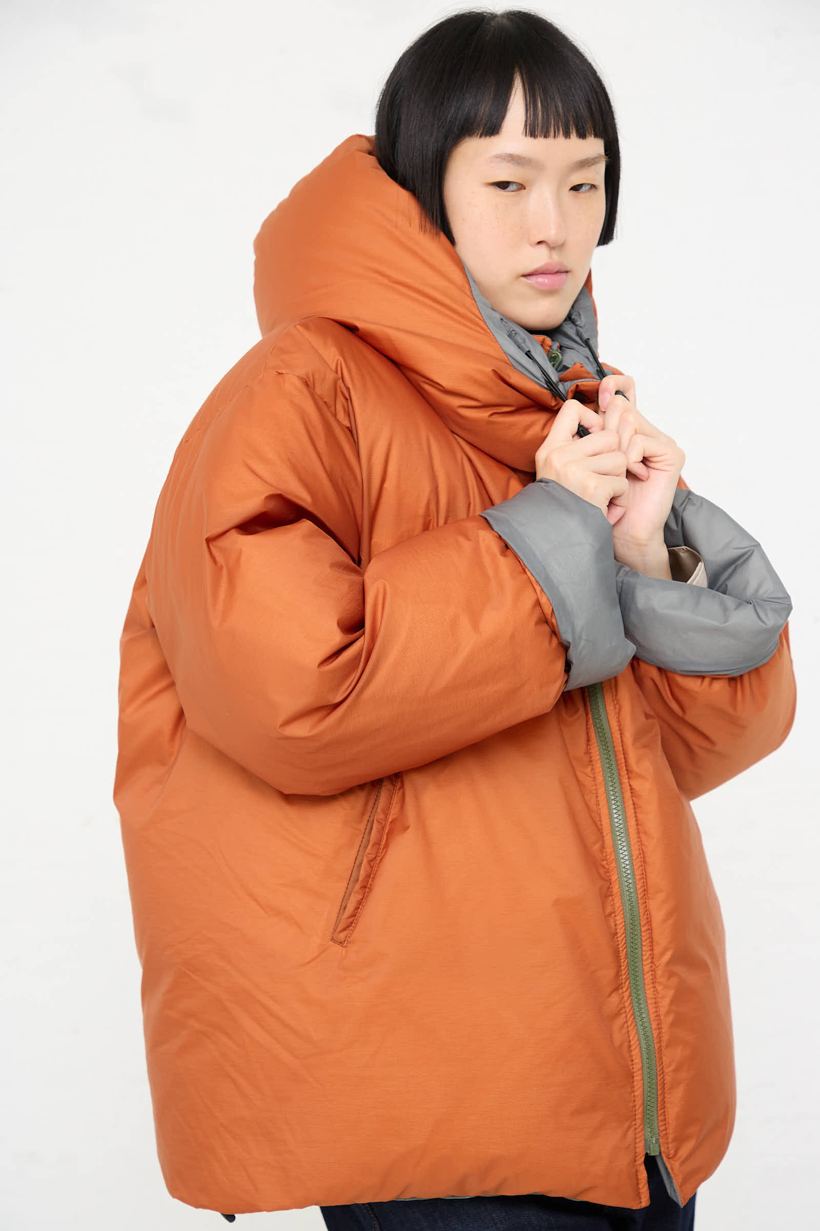 A person wearing the Sofie D'Hoore Down Filled Odna Reversible Jacket in Rust and Sage, an oversized, water-repellent orange puffer with a gray inner lining, stands against a plain white background. They are adjusting the collar with both hands.