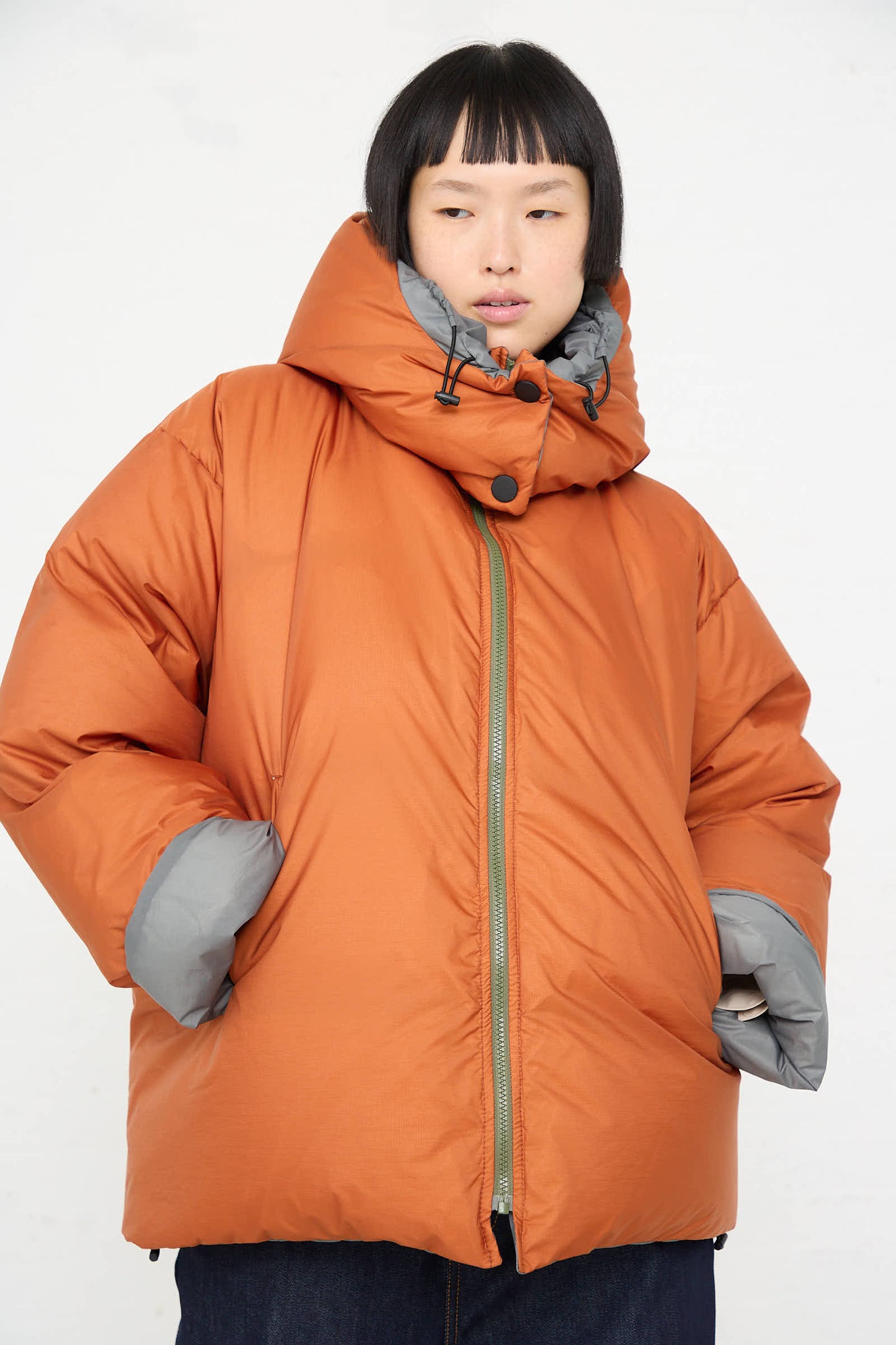 A person wearing a Sofie D'Hoore Down Filled Odna Reversible Jacket in Rust and Sage, with hands in pockets, standing against a plain white background.