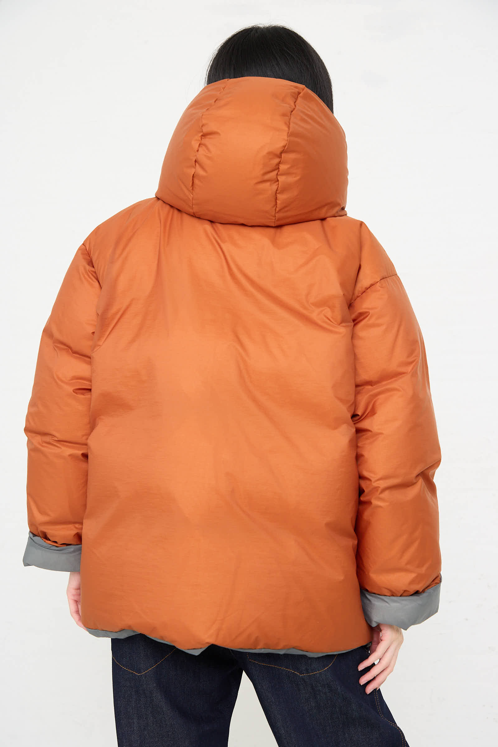 A person with long hair wearing a hooded, water-repellent Down Filled Odna Reversible Jacket in Rust by Sofie D'Hoore and dark jeans stands with their back facing the camera.