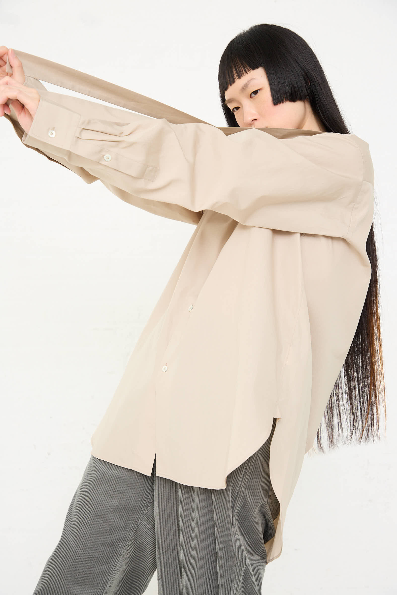 A person with long, straight hair and bangs, wearing a loose-fitting beige Paper Touch Poplin Bow Shirt in Stone by Sofie D'Hoore with a button front closure and gray pants, poses with one arm extended outwards, pulling on the sleeve.