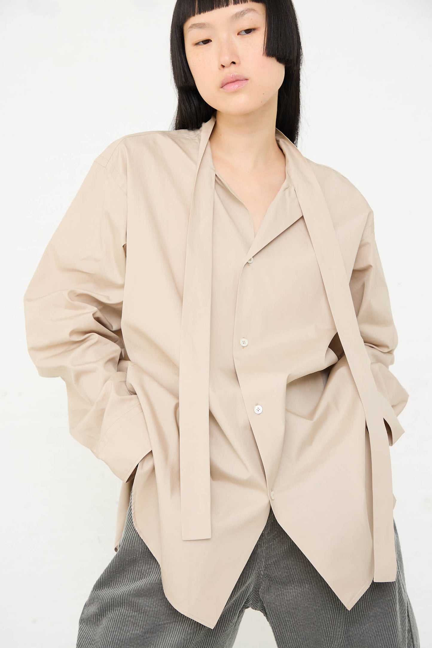 A person is wearing the Sofie D'Hoore Paper Touch Poplin Bow Shirt in Stone, an oversized, long sleeve beige shirt with a button front closure and loose neck ties, paired with gray pants, standing against a plain background.