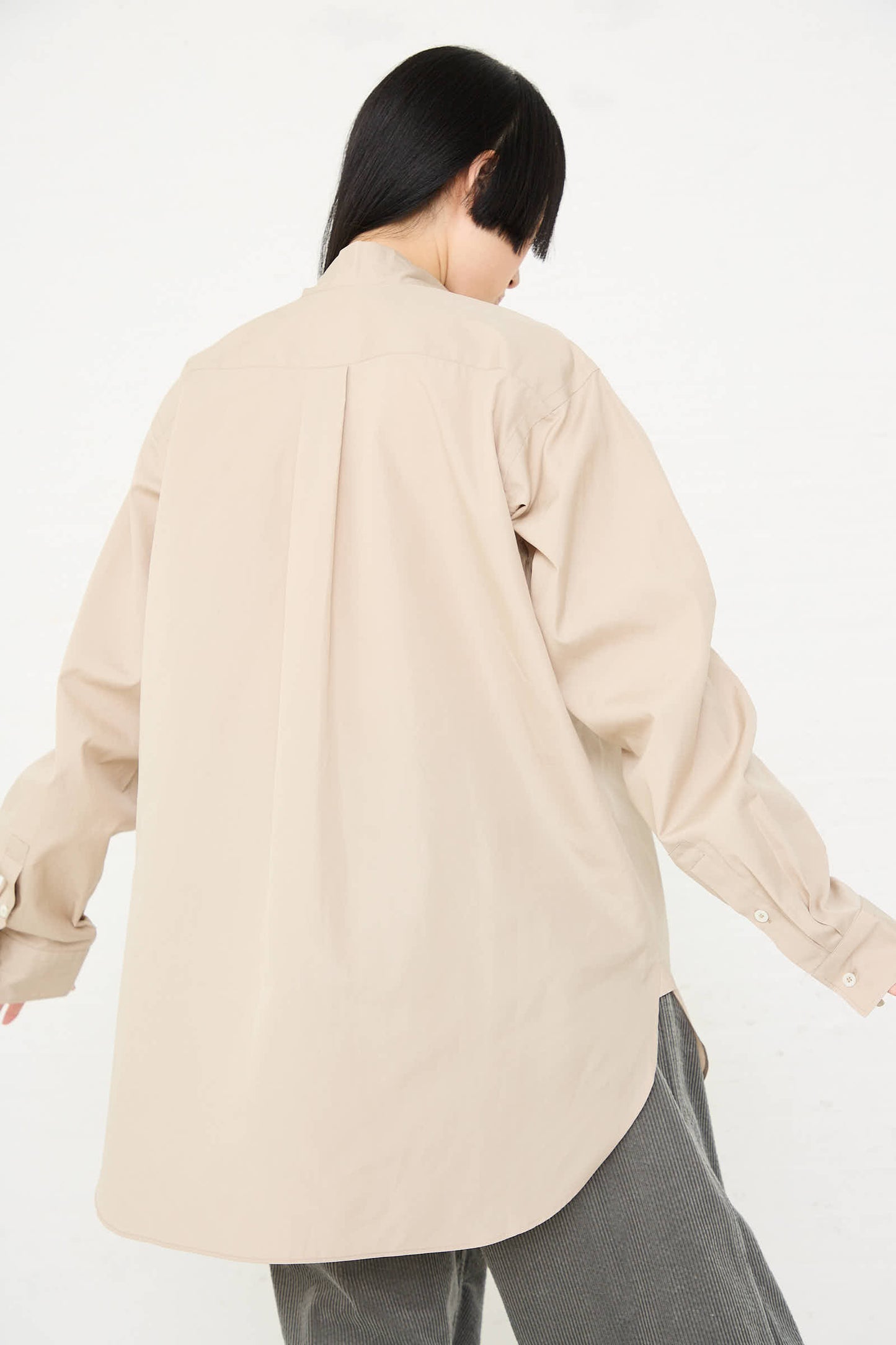 Person stands facing away, wearing a loose-fitting, long-sleeved Paper Touch Poplin Bow Shirt in Stone by Sofie D'Hoore with grey pants. They have shoulder-length black hair. The background is plain white.