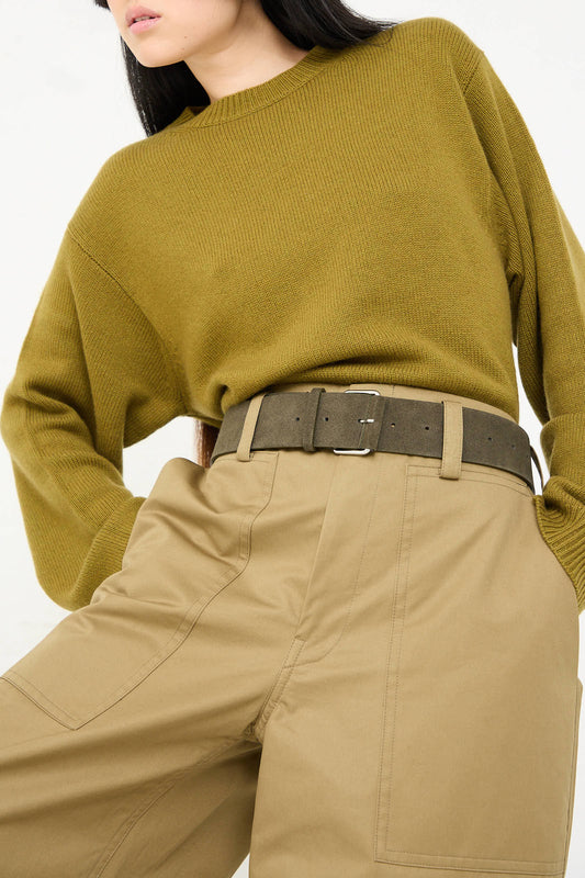 A person wearing an olive green sweater, beige high-waisted trousers with large pockets, and a matching Sofie D'Hoore Suede Leather Ventura Belt in Moss with a silver nickel buckle.