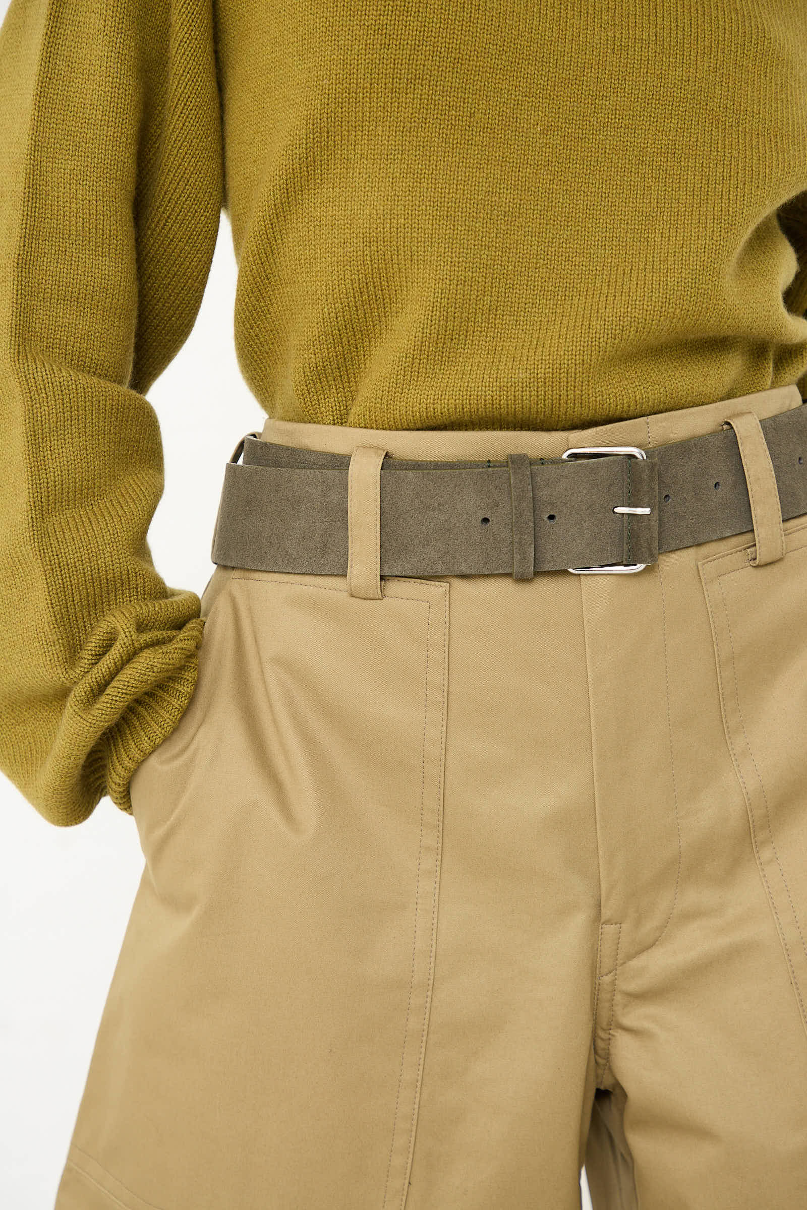 A person wearing a green sweater, beige pants, and the Sofie D'Hoore Suede Leather Ventura Belt in Moss with a silver nickel buckle.