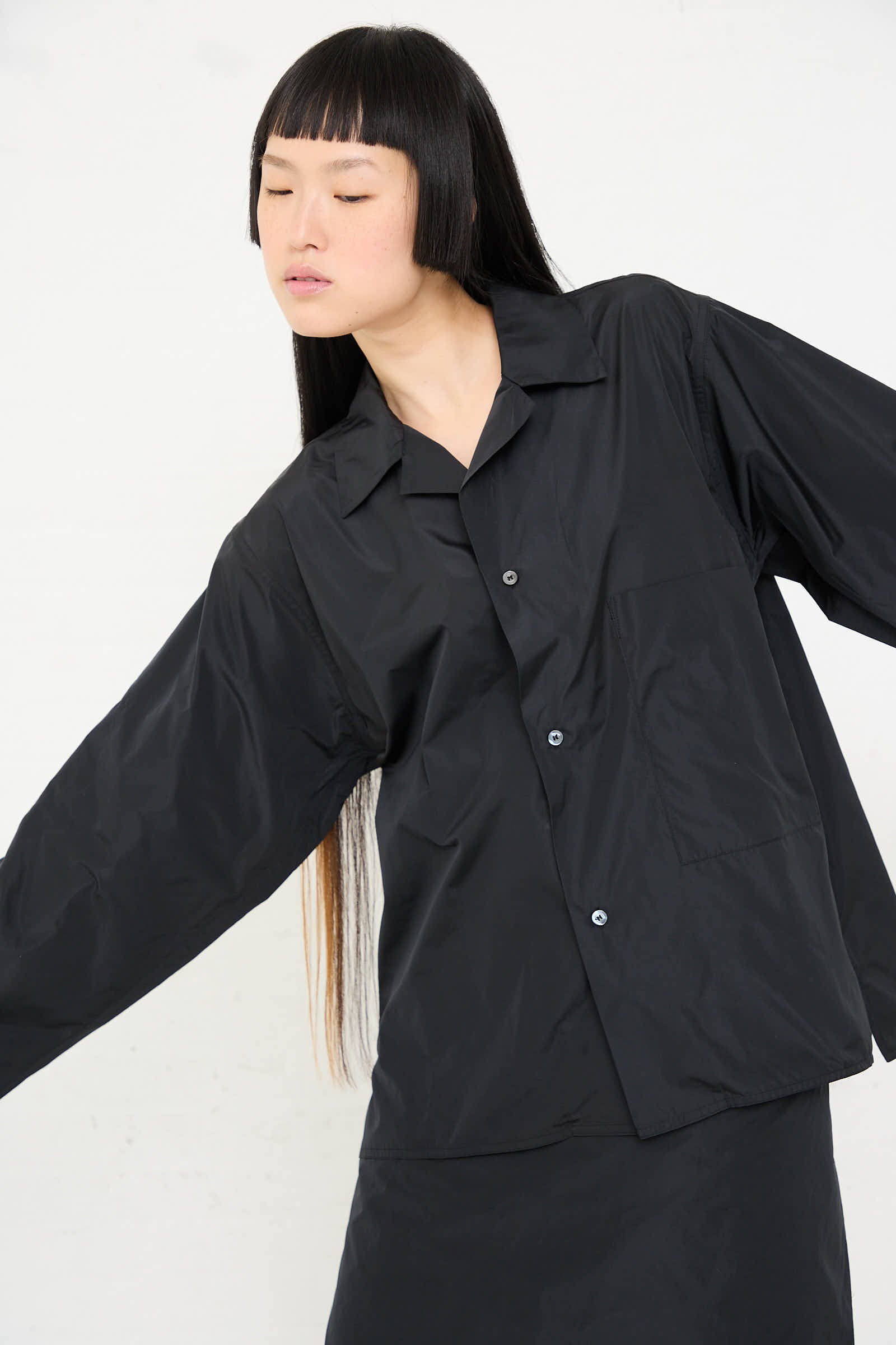 Person with long hair and bangs wearing the loose-fitting, black "Taffeta Barry Shirt with Patch Pocket" by Sofie D'Hoore, crafted from lightweight polyester, paired with black pants and posing with arms slightly outstretched.