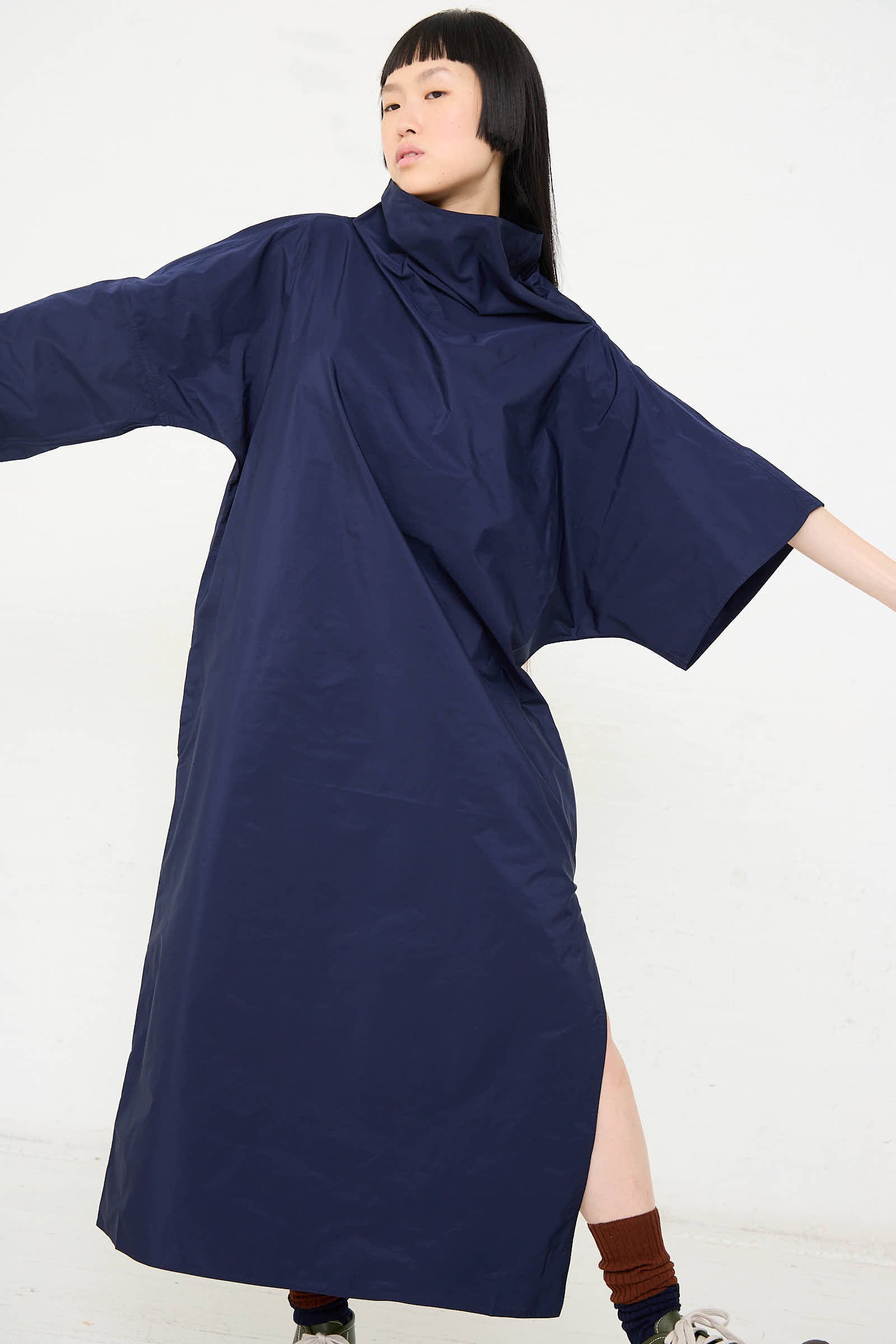 A person wearing the Taffeta Dee Dee Dress in Touareg by Sofie D'Hoore, featuring an oversized fit, long sleeves, and a side slit, paired with dark socks and sneakers. The individual is extending their arms with a neutral expression against a white background.