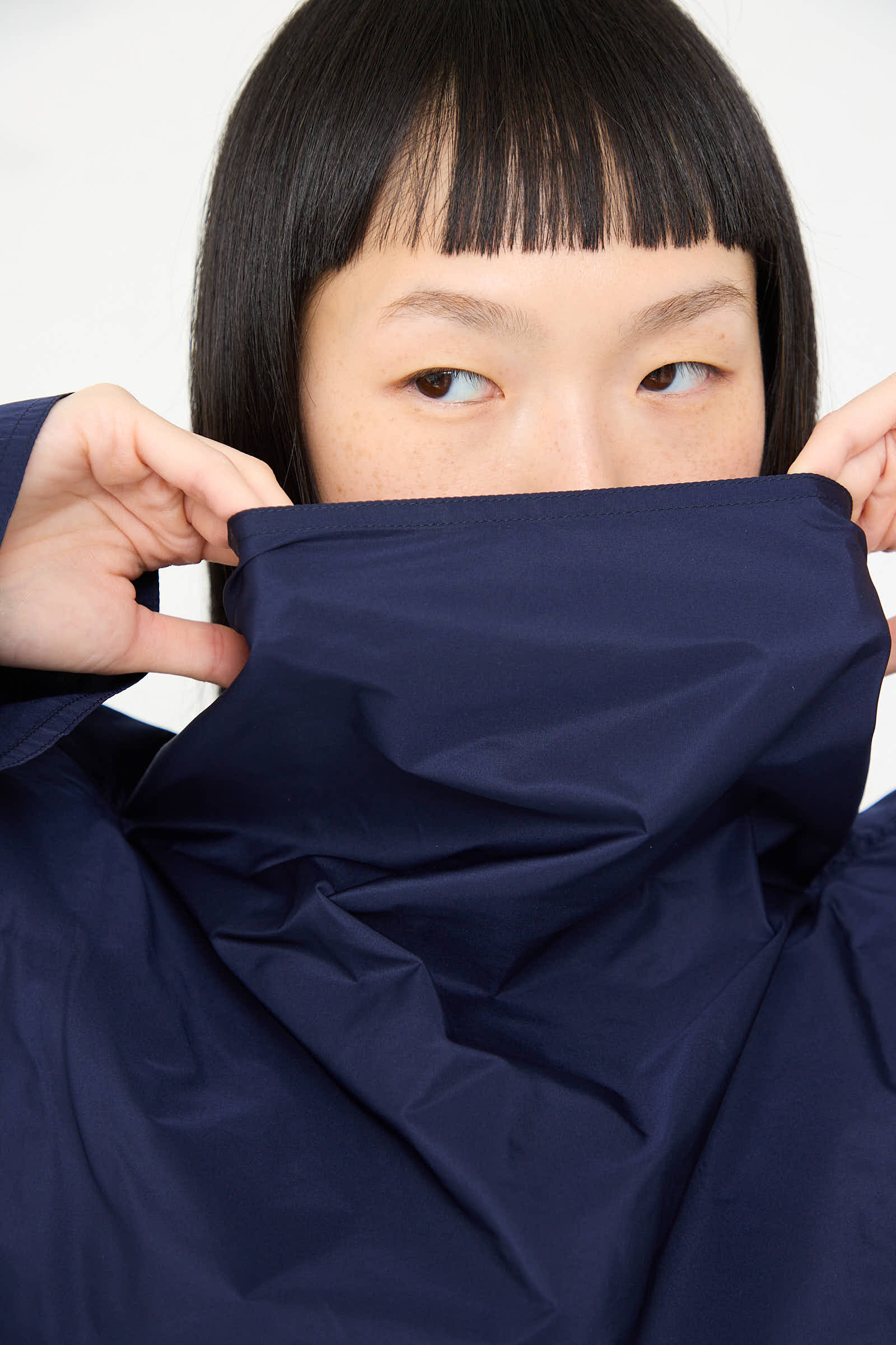 A person with straight black hair covering the lower half of their face with a navy blue fabric, looking to the side, wears the Taffeta Dee Dee Dress in Touareg by Sofie D'Hoore.