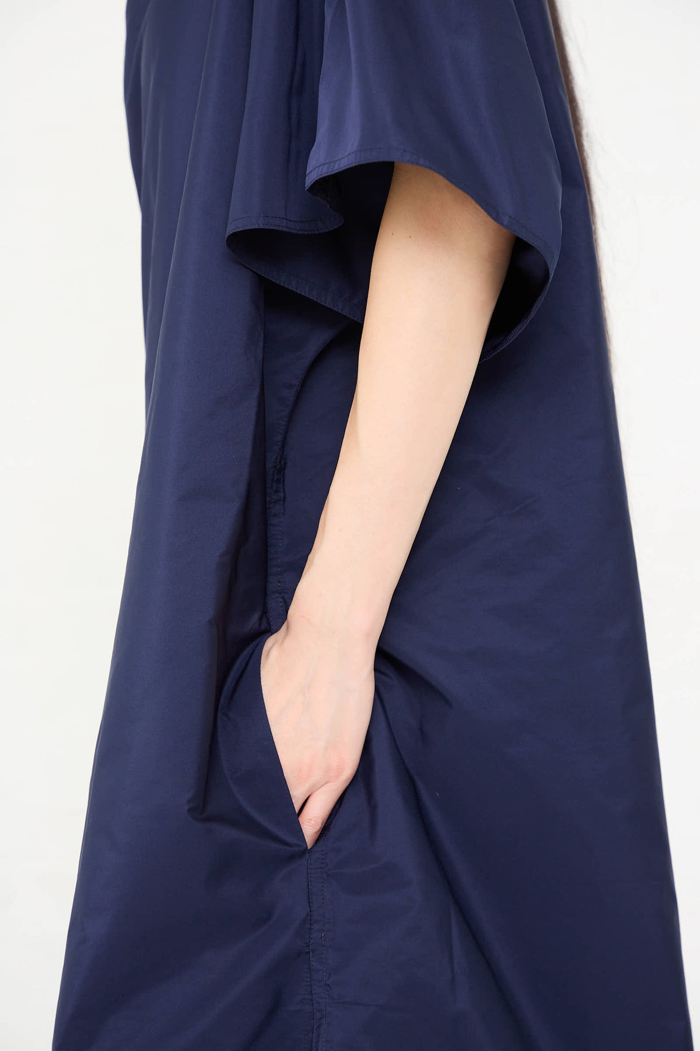 Close-up of a person wearing the Taffeta Dee Dee Dress in Touareg by Sofie D'Hoore, a navy blue, short-sleeved, oversized dress with one hand in a side pocket.