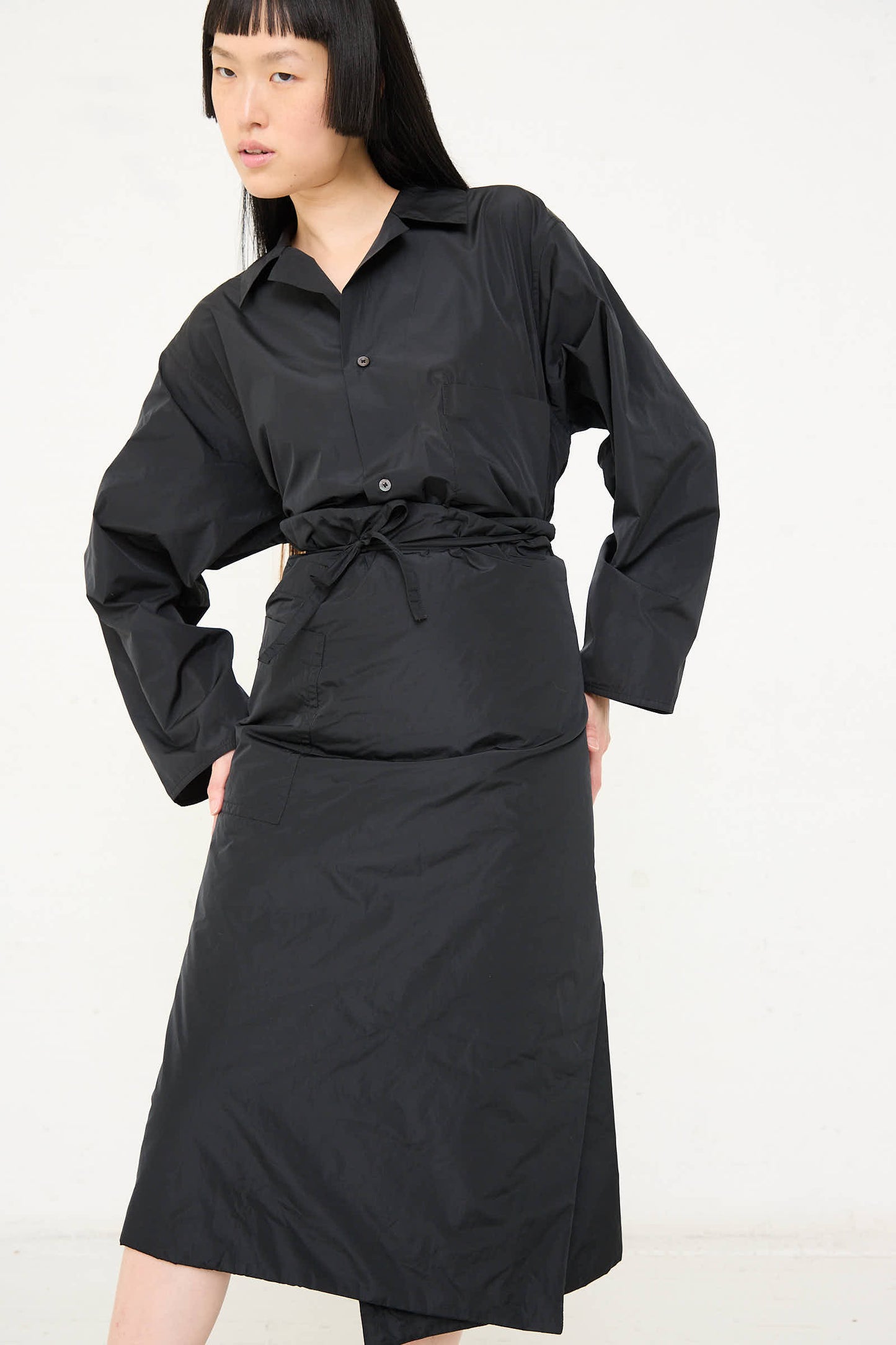 Person wearing a long black button-up shirt and the Taffeta Long Padded Wrap Sill Skirt in Black from Sofie D'Hoore stands against a plain white background.