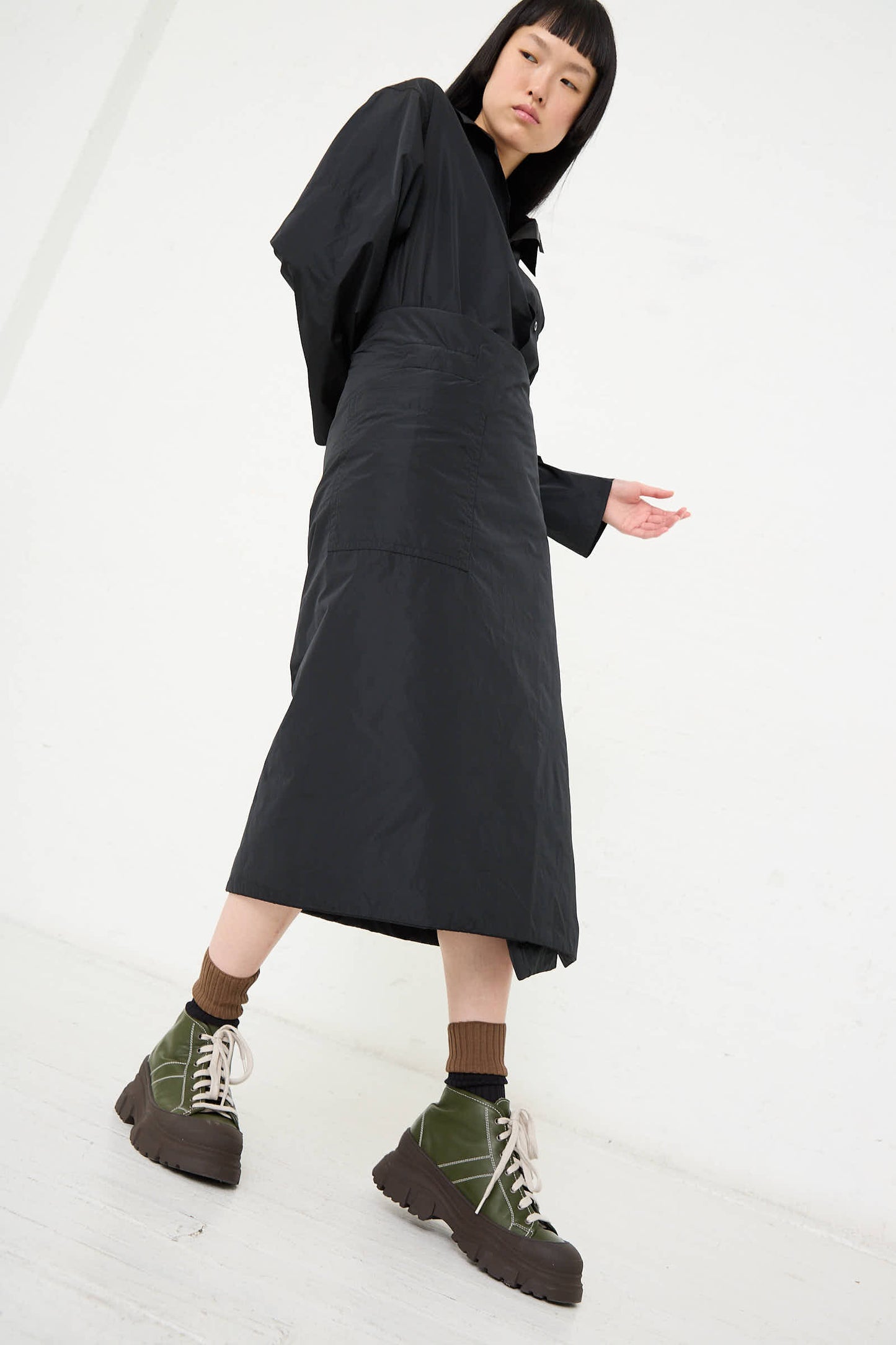 A person wearing a Sofie D'Hoore Taffeta Long Padded Wrap Sill Skirt in Black, brown socks, and green chunky sneakers stands against a white backdrop.