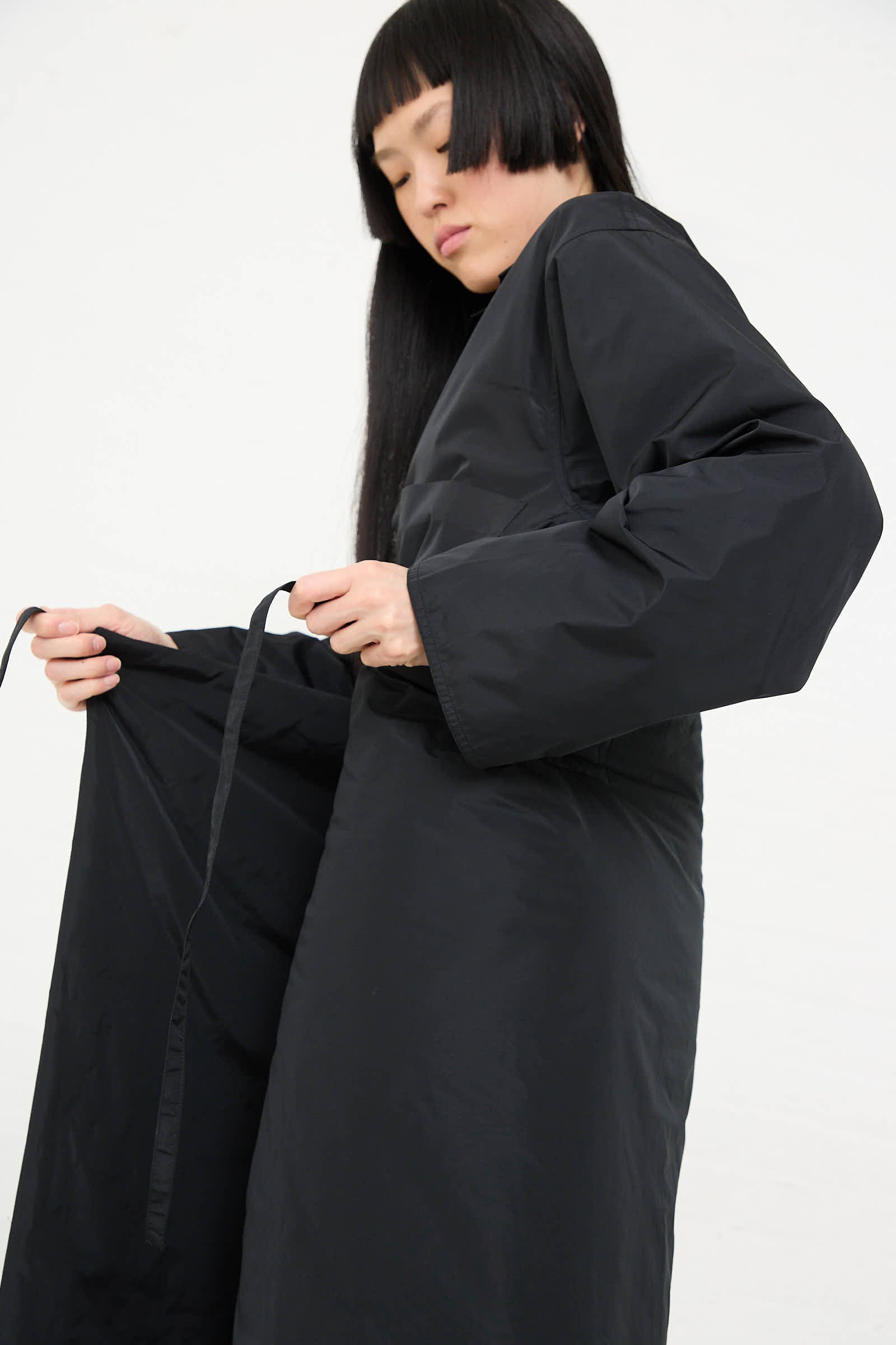 Person with long black hair ties a black robe around their waist against a plain background, complementing the look with a relaxed fit Taffeta Long Padded Wrap Sill Skirt in Black by Sofie D'Hoore.