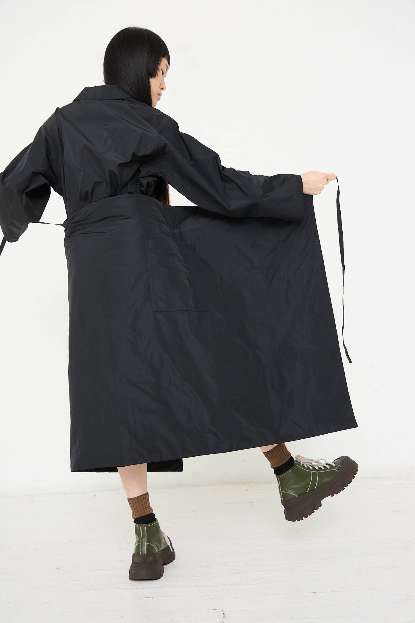 Person in a black Taffeta Long Padded Wrap Sill Skirt by Sofie D'Hoore, tying the belt of the skirt, standing on a light-colored floor with relaxed fit olive green boots.