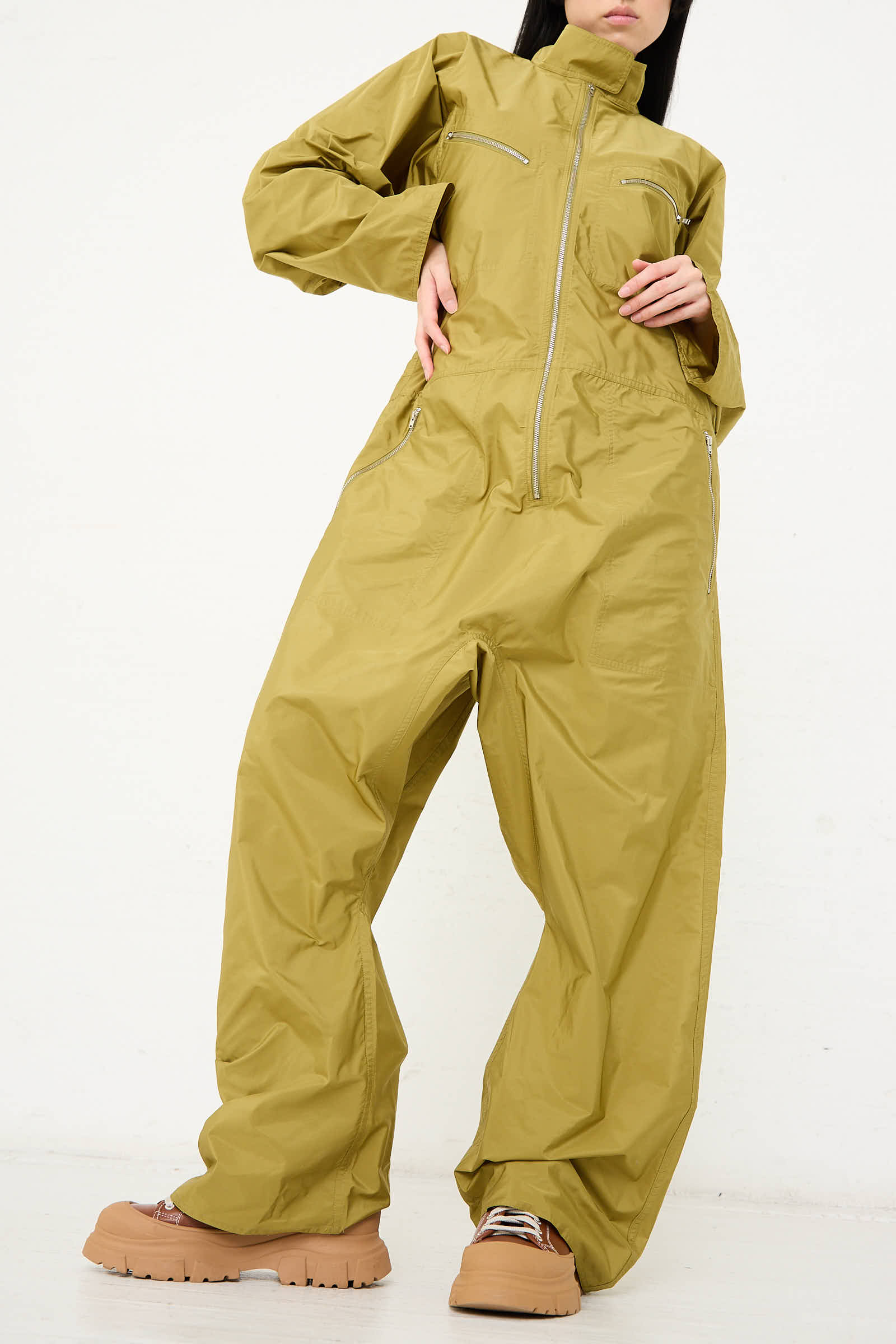 Person wearing an oversized olive Taffeta Plateau Jumpsuit by Sofie D'Hoore with a front zip closure and tan and pink platform sneakers, standing against a plain white background.