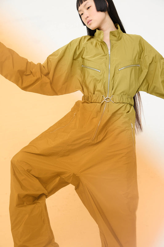 A person with long hair, wearing the Taffeta Plateau Jumpsuit in Olive by Sofie D'Hoore, poses against a light background with one arm raised and the other bent at the elbow.