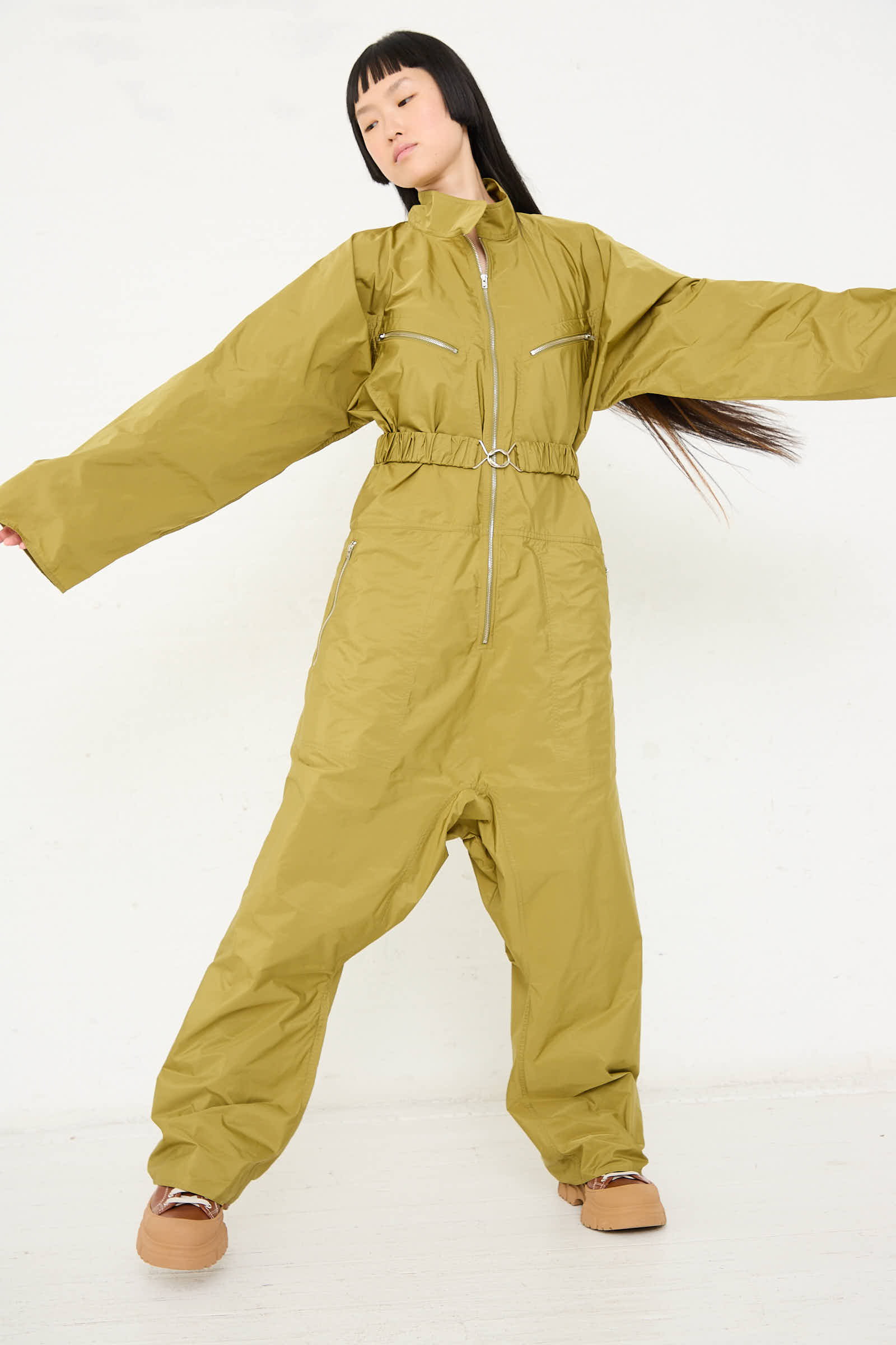 A person stands with arms outstretched, wearing the oversized Taffeta Plateau Jumpsuit in Olive from Sofie D'Hoore, featuring a front zip closure and beige shoes, against a plain white background.