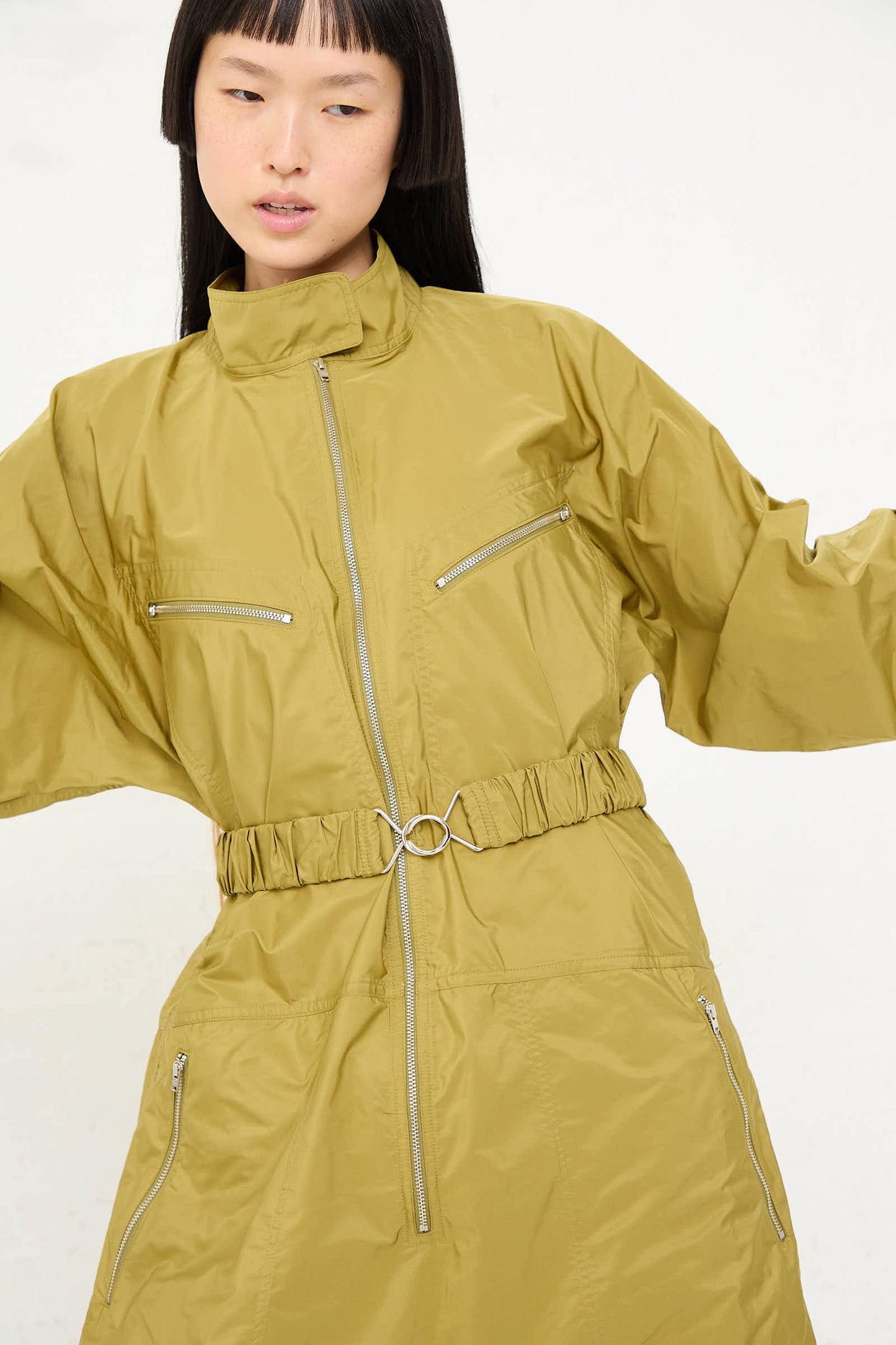 A person wearing the Taffeta Plateau Jumpsuit in Olive by Sofie D'Hoore, a stylish oversized polyester jumpsuit featuring a belted waist and multiple front zip closures, is posing against a plain background.