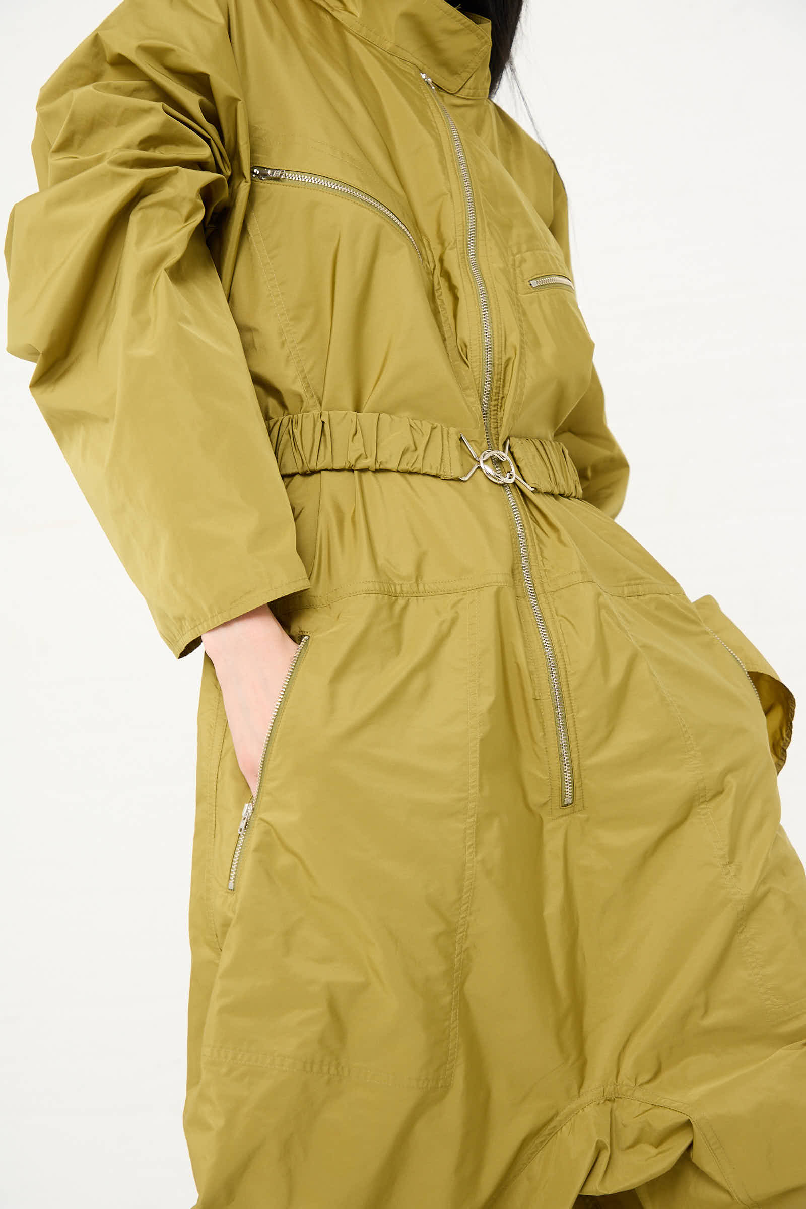 A person wearing the Sofie D'Hoore Taffeta Plateau Jumpsuit in Olive, featuring long sleeves, a front zip closure, and an elasticated waist.
