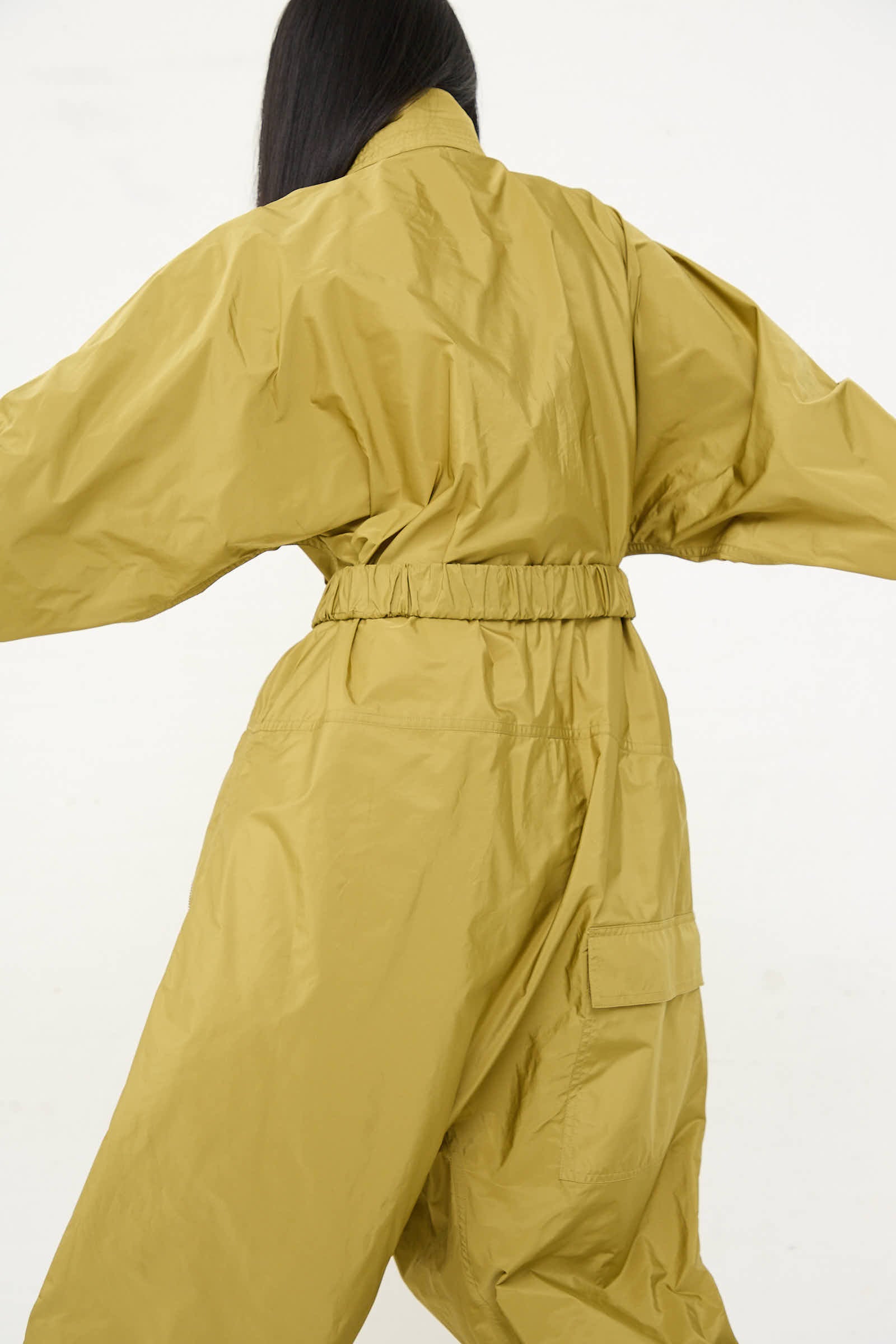 Person wearing an oversized olive jumpsuit with front zip closure, facing away from the camera with arms slightly raised. The jumpsuit is the Taffeta Plateau Jumpsuit by Sofie D'Hoore.