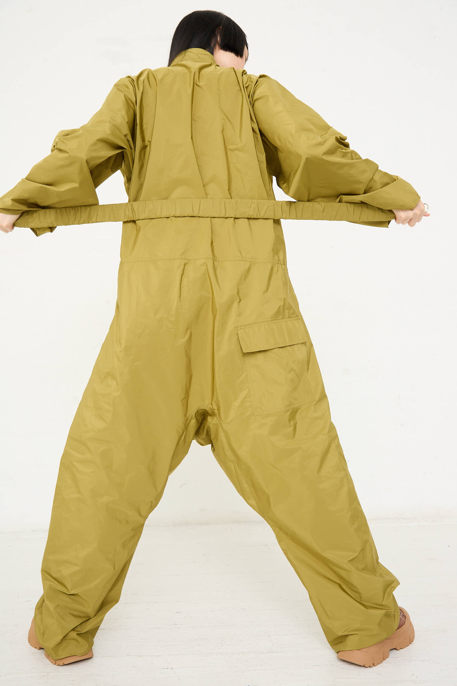 Person in an oversized Taffeta Plateau Jumpsuit in Olive by Sofie D'Hoore, featuring a front zip closure, facing away, holding the suit's belt with both hands, and standing against a plain white background.