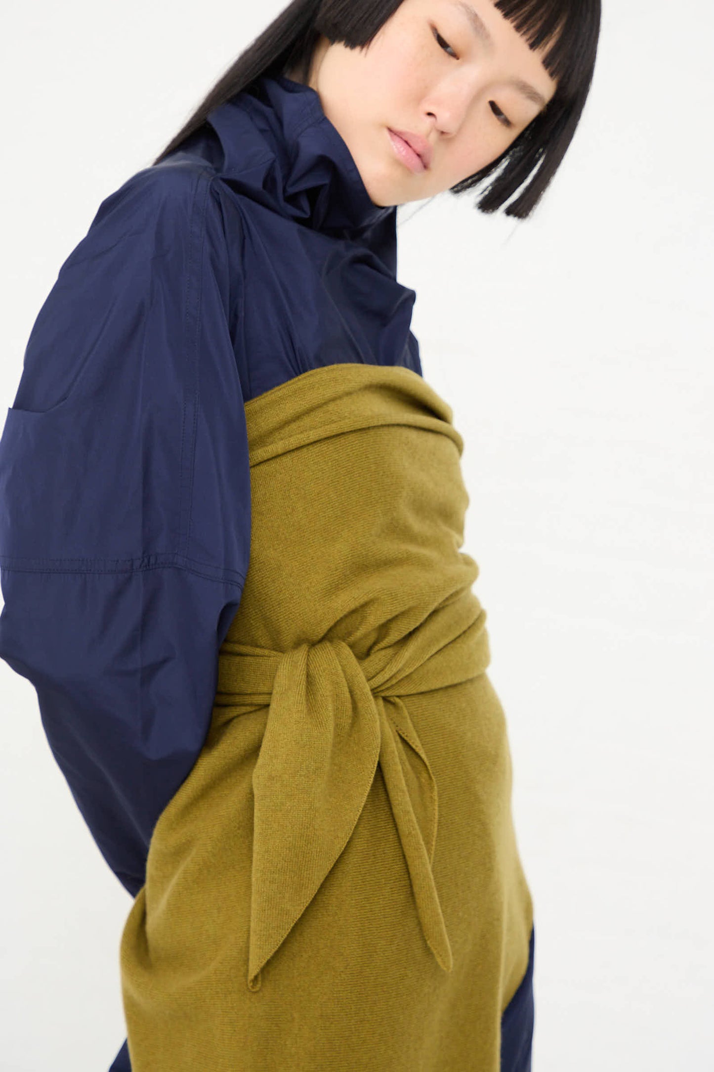 Person wearing a blue jacket with the Sofie D'Hoore Wool Cashmere Astra Scarf in Olive, tied at the waist, looking down.