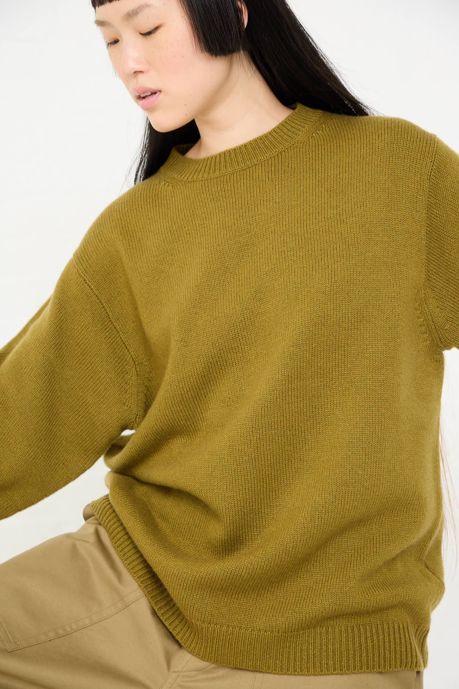 A person with long, dark hair wearing a loose-fitting Wool Cashmere Moody Sweater in Olive by Sofie D'Hoore and beige pants looks downwards.