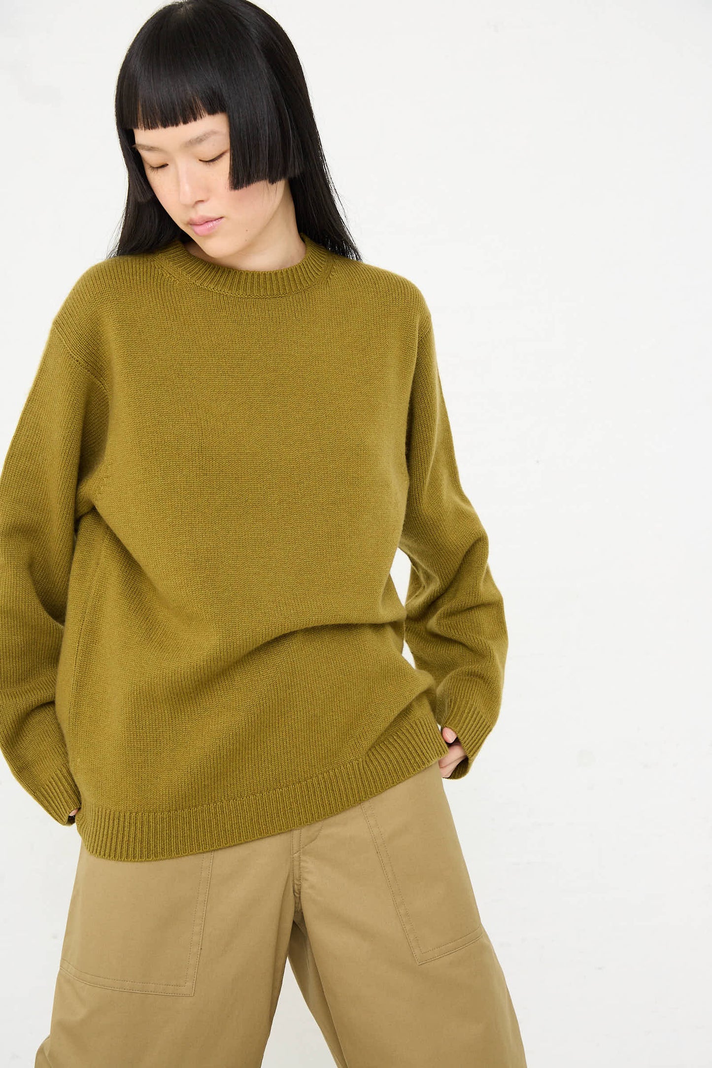 A person with long black hair wearing a Sofie D'Hoore Wool Cashmere Moody Sweater in Olive and khaki pants stands against a plain background.