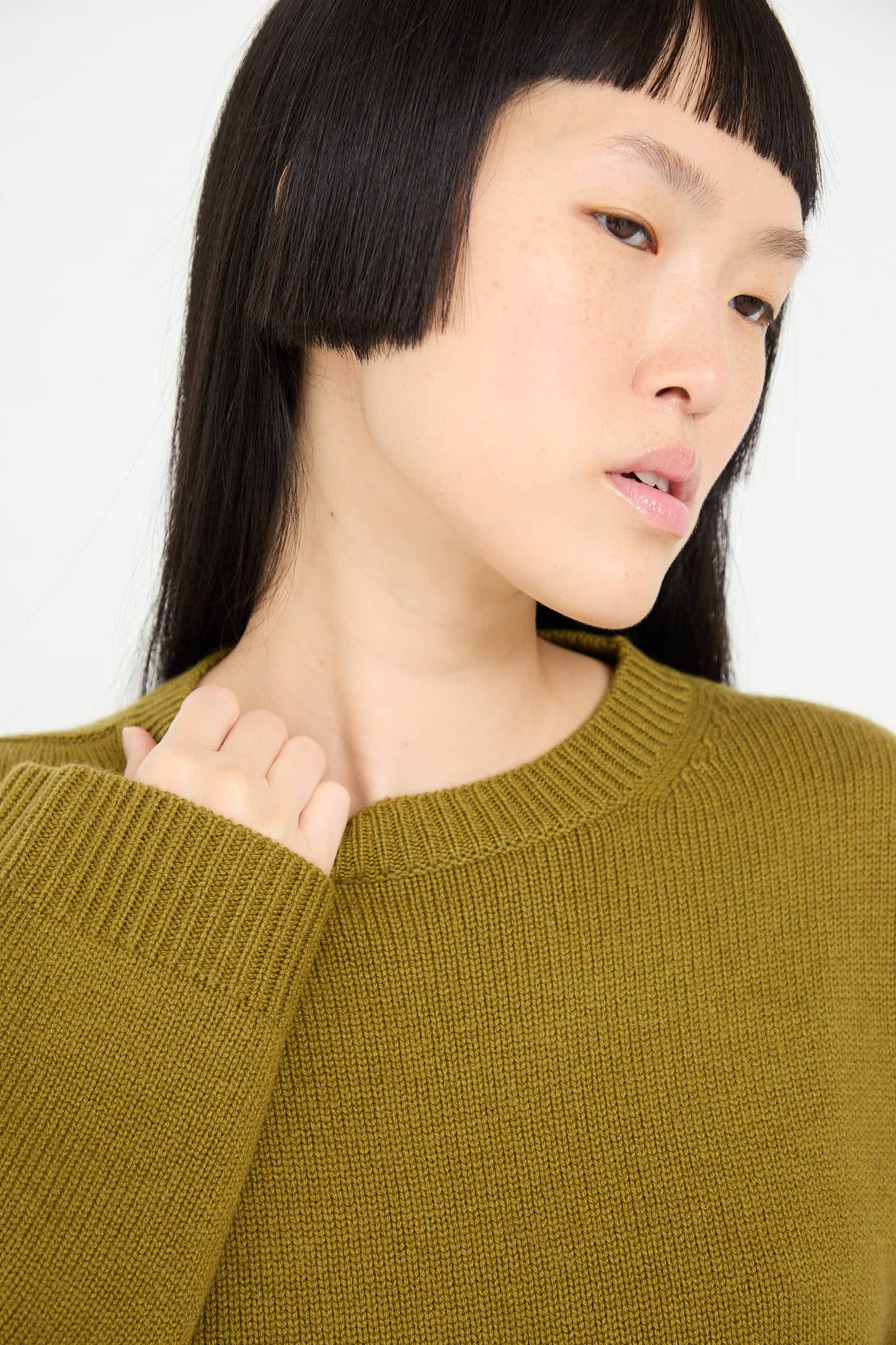 A person with long black hair and bangs looks off to the side while wearing the Wool Cashmere Moody Sweater in Olive from Sofie D'Hoore. One hand is touching the luxurious wool-cashmere blend near the shoulder.