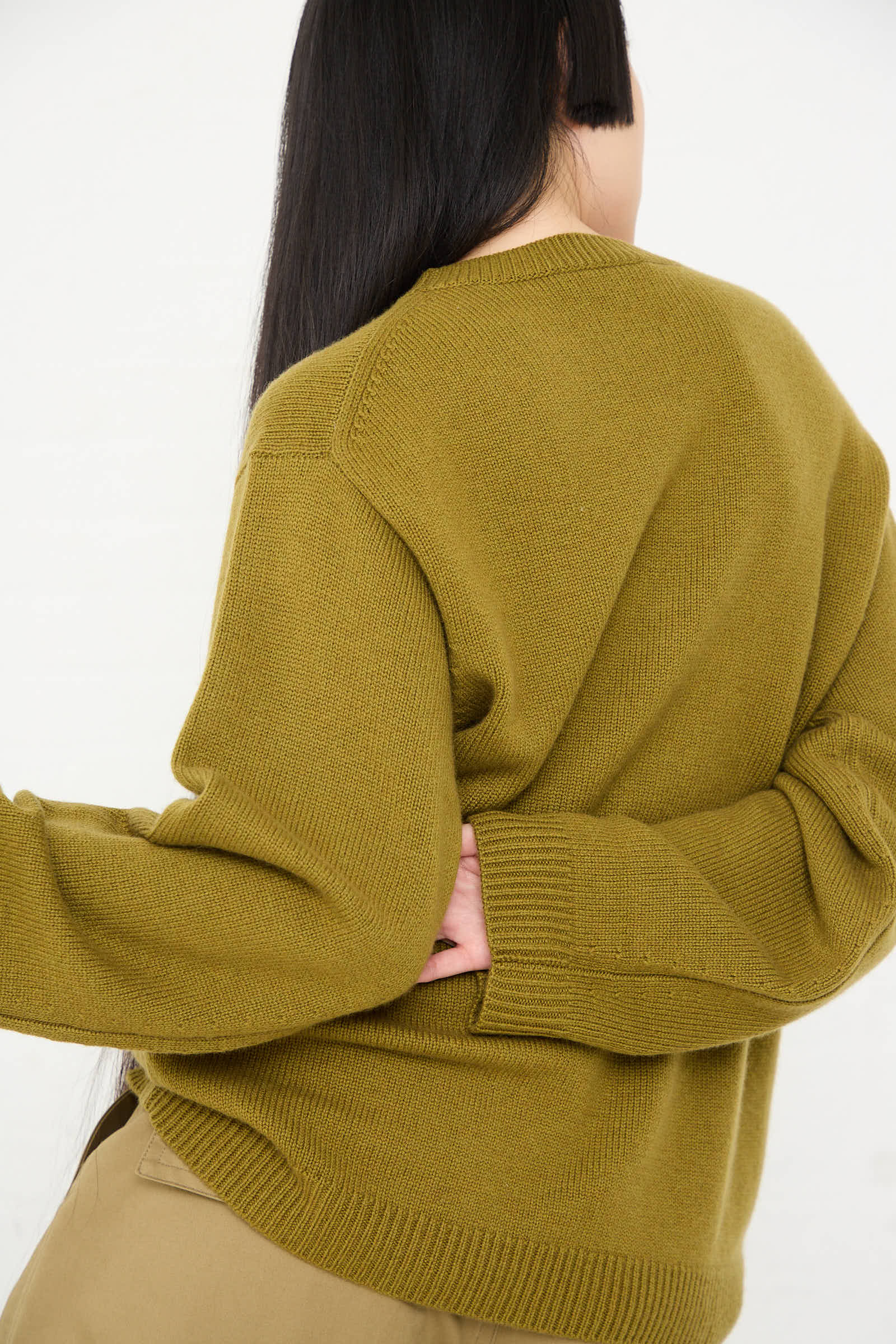 A person with long black hair, seen from the back, is wearing the Sofie D'Hoore Wool Cashmere Moody Sweater in Olive and beige pants against a plain white background.