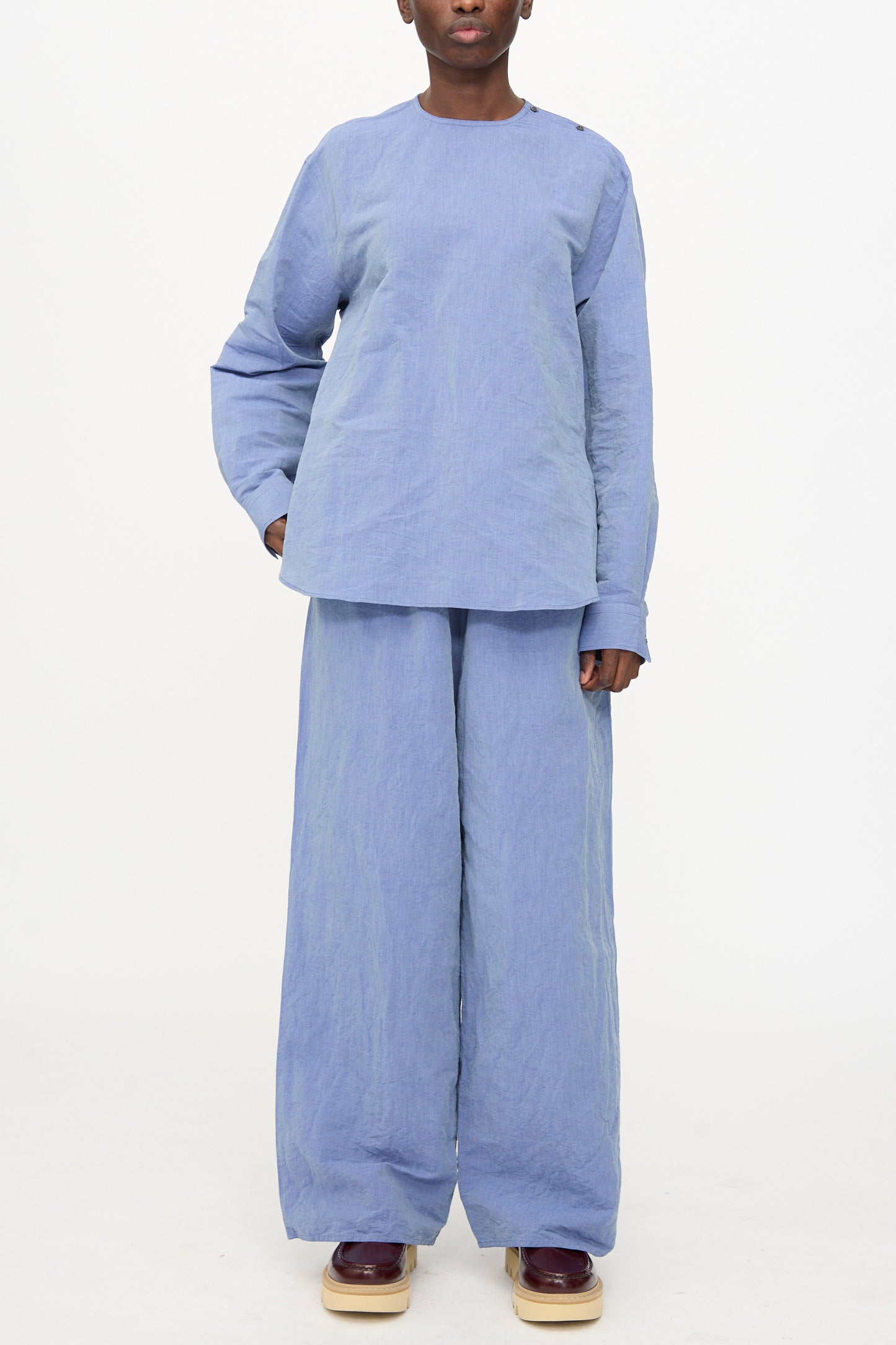 Person standing against a plain background, wearing the Sofie D'Hoore Barel Crewneck in Blue Jeans with matching wide-leg pants and platform shoes.