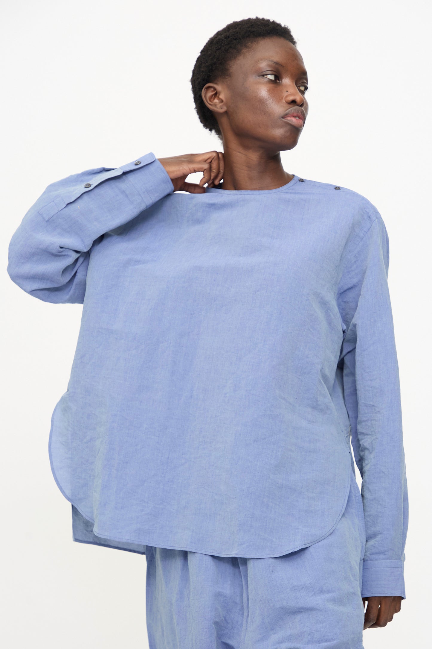 A person wearing the Sofie D'Hoore Barel Crewneck in Blue Jeans, styled with matching pants, looks to the side and touches their neck against a plain background.