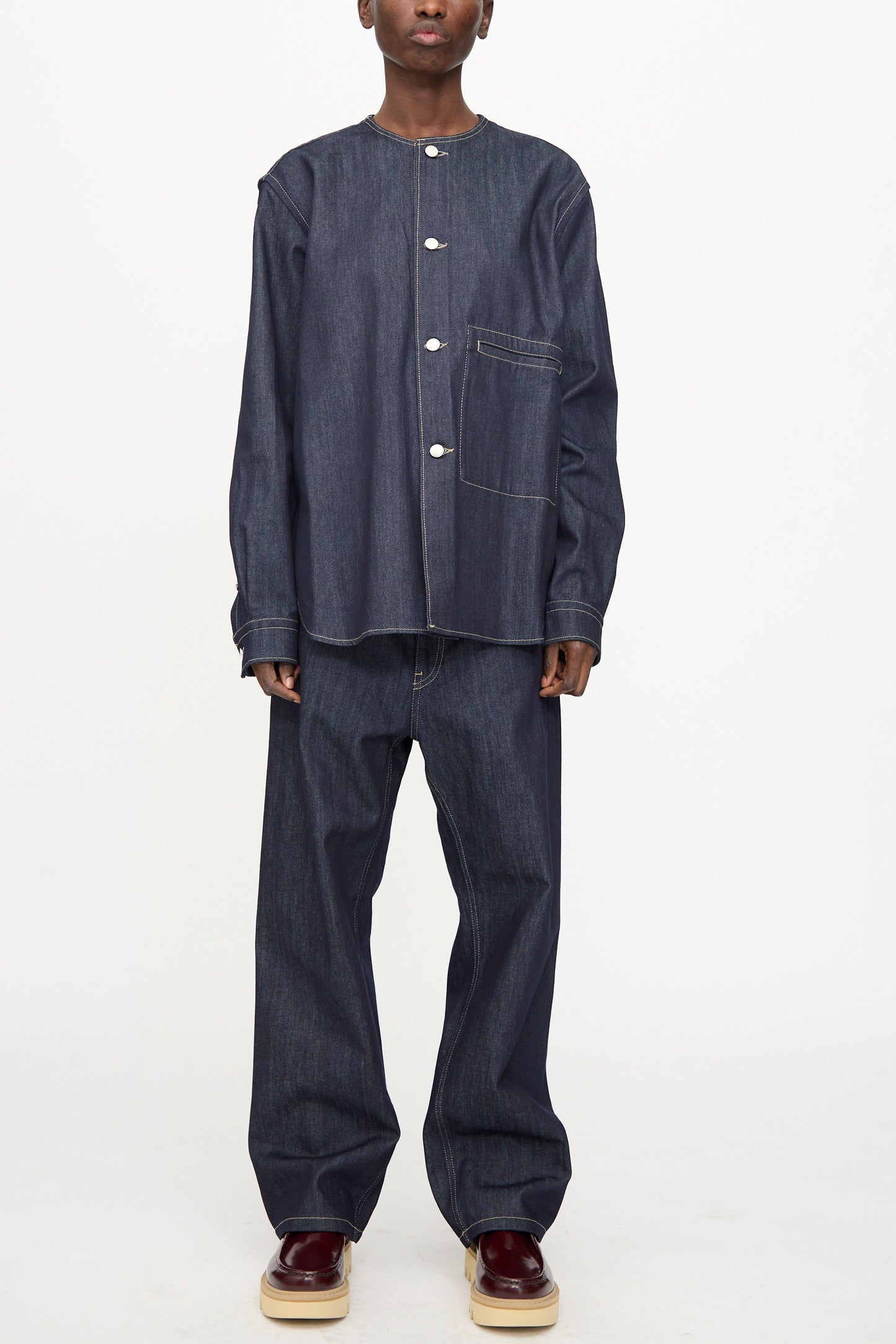 A person wearing the Sofie D'Hoore Barna Shirt in Midnight—a relaxed fit, long sleeve raw denim top with a front pocket—paired with matching wide-leg denim pants and burgundy platform shoes, stands against a plain white background.